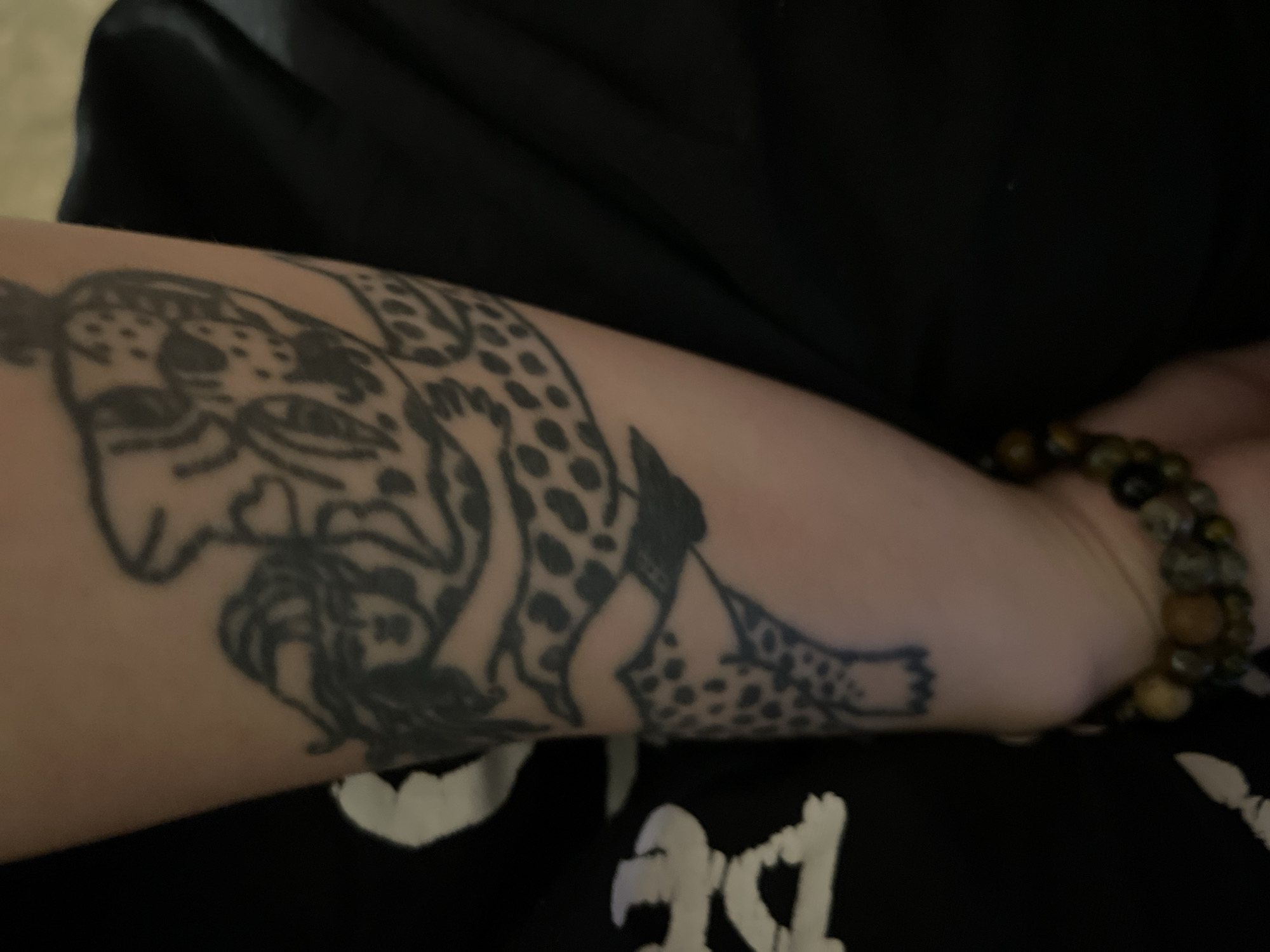 The image displays a close-up of a person's forearm and wrist, featuring a prominent tattoo and a bracelet. The skin tone is fair.
The main subject is a large, black outline tattoo covering a significant portion of the forearm. The tattoo depicts a stylized spotted big cat, possibly a leopard or jaguar, in a dynamic pose. Its head is oriented towards the left side of the frame, showing distinct eyes, a nose, and a mouth. The creature's body is adorned with numerous irregular black spots, and its form seems to intertwine with what appears to be a human-like figure or face, specifically a woman with flowing dark hair, visible within the cat's design. The design extends from the upper forearm down towards the wrist.
On the wrist, a bracelet made of multiple round, polished beads is visible. The beads vary in color from light brown to dark brown and black, suggesting natural stones like tiger's eye.
The arm is positioned against a background of dark, possibly black, fabric, likely part of the clothing worn by the person. A section of black fabric with abstract white shapes or stylized graphic elements is visible at the bottom of the image, possibly from a t-shirt or other garment.
The lighting suggests an indoor setting. No specific location cues are visible within the image itself, but the provided context places it in Picassent, Spain. No discernible text is clearly visible in the tattoo or on the clothing.