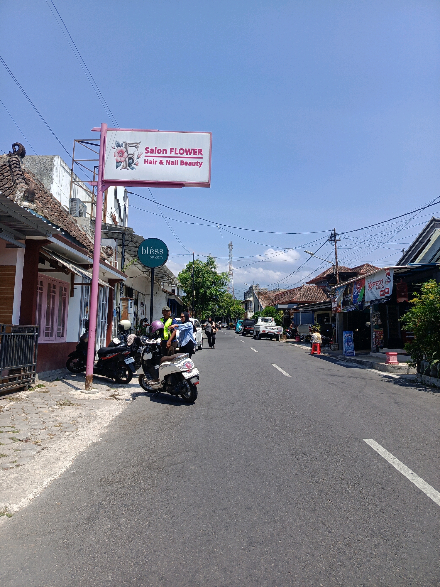 The image depicts a street scene in Kebonagung, Indonesia, on a clear, sunny day. The perspective is from the roadside, looking down a moderately trafficked road lined with commercial establishments. A prominent sign for "Salon FLOWER Hair & Nail Beauty" is visible, with a pink floral design. Next to it, a circular green sign reads "bless bakery." Several motorcycles are parked along the sidewalk, indicating common modes of transportation. People are seen walking and interacting on the street, including two individuals near the motorcycles who appear to be conversing. Buildings on either side of the road showcase local architecture, with tiled roofs and open storefronts. Utility poles with a complex network of wires crisscross the sky, a common sight in urban Indonesian landscapes. Further down the road, more shops and a white pickup truck are visible, suggesting a commercial district. The overall impression is one of a typical day in a bustling Indonesian town.