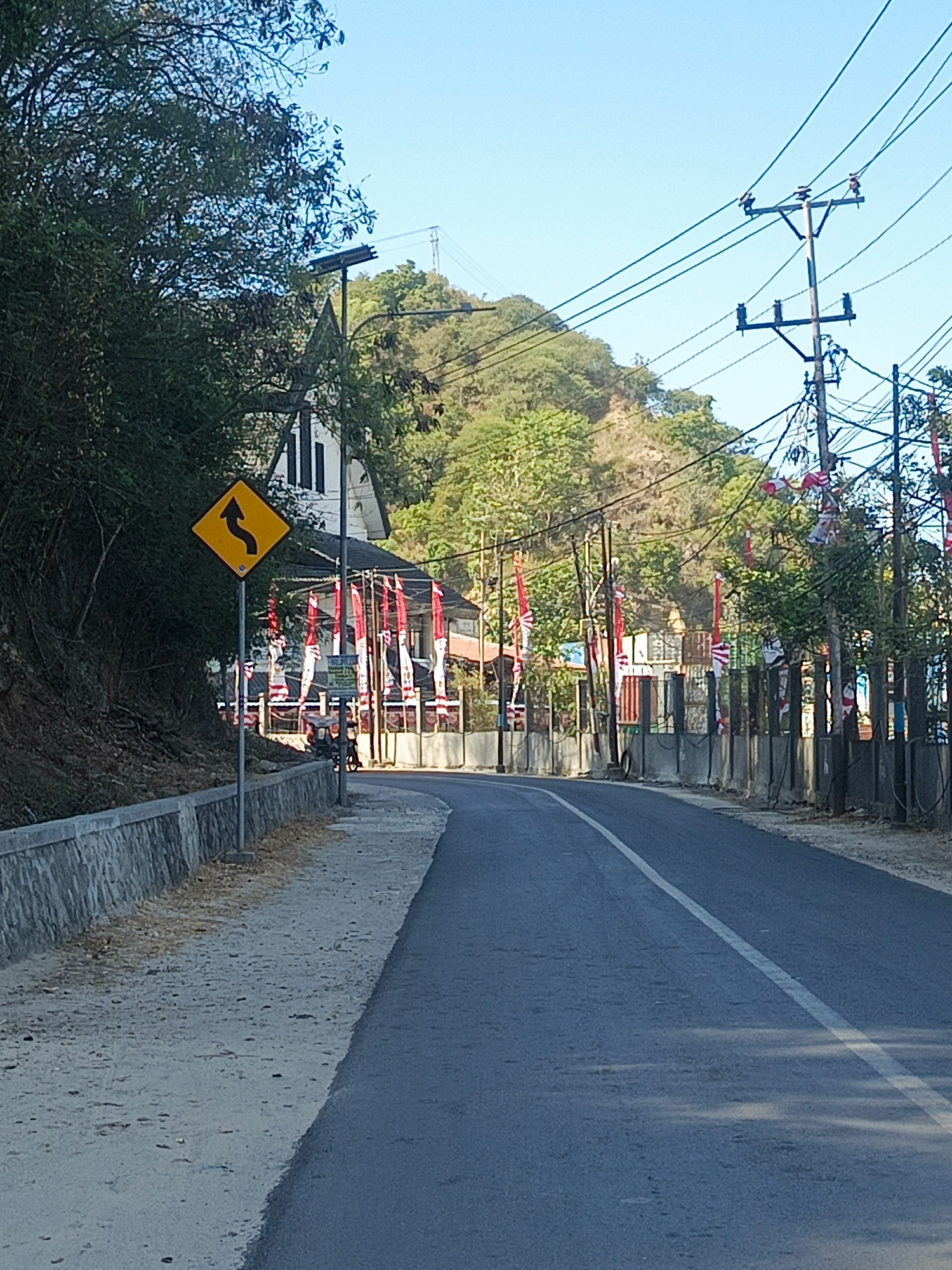 The image depicts a paved road curving to the right, with a sidewalk and a stone retaining wall on the left. A yellow diamond-shaped sign with a black arrow indicating a winding road is prominently displayed on the sidewalk. Beyond the sidewalk, a white building with dark trim is visible, adorned with numerous red and white flags, suggesting a national celebration. Overhead, power lines stretch across a clear blue sky, supported by utility poles. Trees and a hillside covered in vegetation form the backdrop. The scene appears to be a street in Gorontalo, Indonesia, likely during a patriotic event, given the prevalence of the national flag. No individuals are clearly identifiable or engaged in any specific actions within the frame. The environment is rural or semi-rural, characterized by natural vegetation and infrastructure.