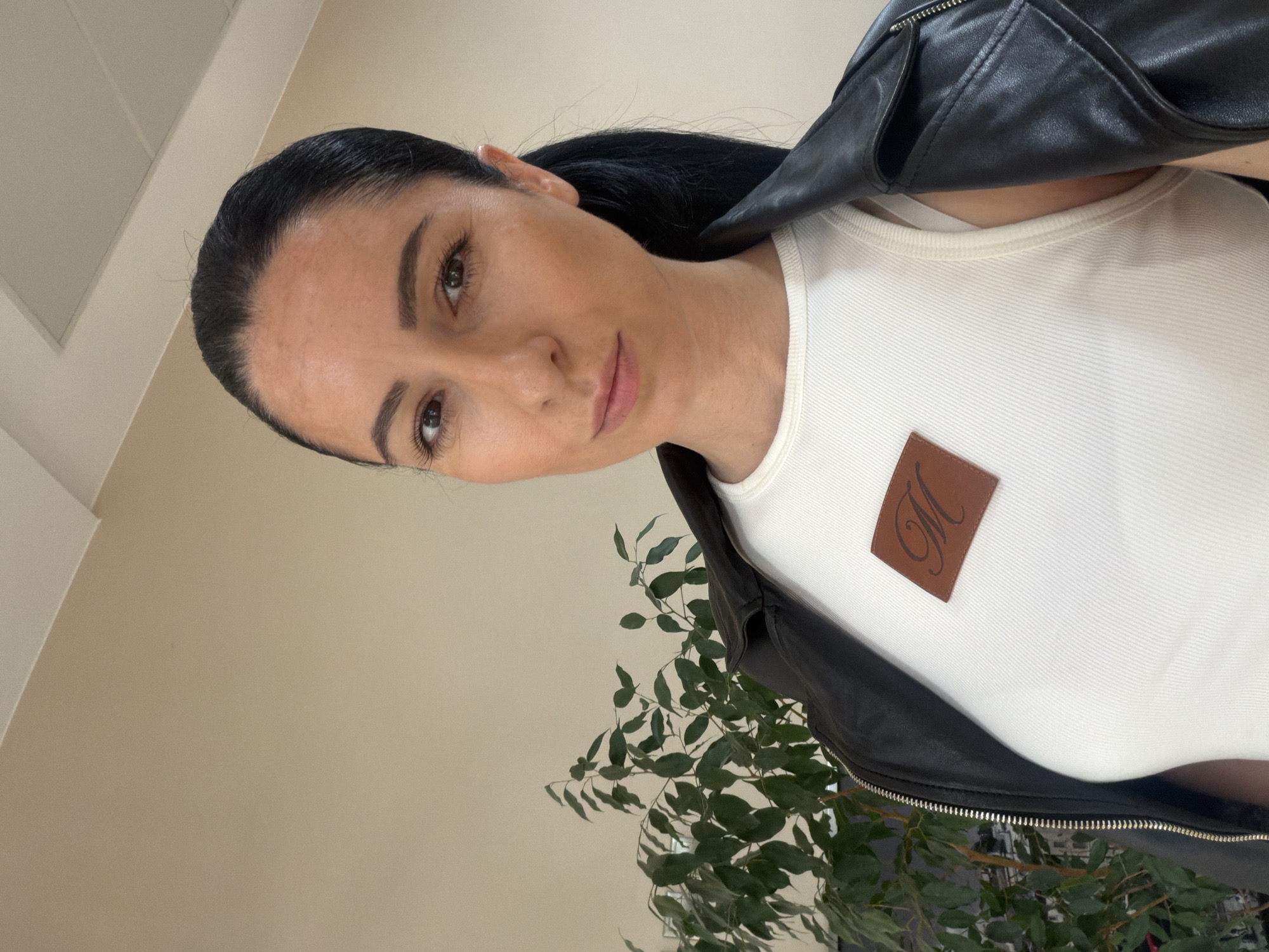 The image depicts a woman with dark hair, wearing a white top with a brown square patch on the left side, and a black leather jacket. A potted plant with green leaves is visible in the background. The location is not specified.