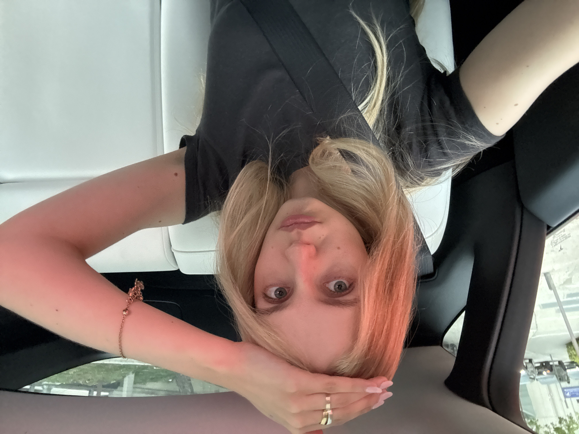 In a car interior, a young Caucasian woman with blonde hair, pale skin, and light gray eyes is taking a selfie. She is seated in the back of a vehicle, wearing a dark gray t-shirt and a seatbelt. Her right hand is raised, touching her forehead, adorned with a gold ring on her index finger. A rose gold bracelet with small charms is visible on her right wrist. The car interior features white leather seats. Outside the car, through the window, a street scene is visible with parked cars and a walkway. The lighting suggests an outdoor setting, possibly in a city, with a warm, reddish hue cast over the subject, indicating a sunset or artificial lighting. The image appears to be taken in Miami, Florida, United States of America.