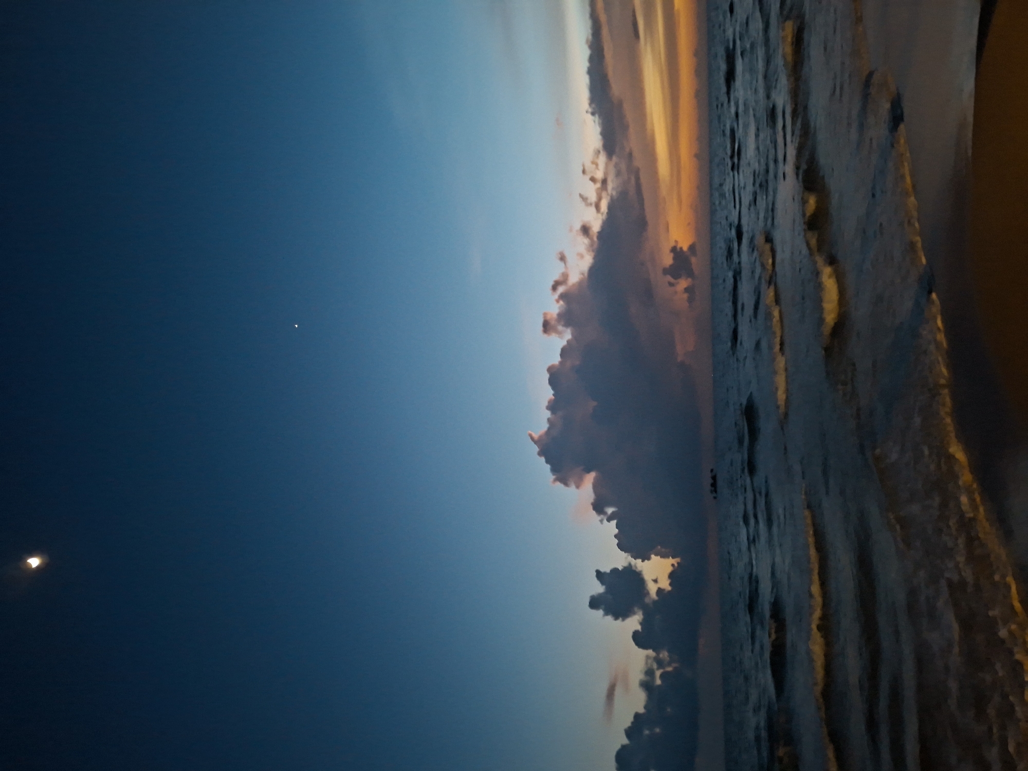 The image depicts a twilight scene at an unknown beach location. The sky is a deep blue, transitioning to hues of orange and yellow near the horizon, indicative of sunset or sunrise. A crescent moon is visible in the upper left portion of the sky, along with a small, bright star. Dark, textured clouds are scattered across the sky, some illuminated by the fading light. The ocean surface is calm with gentle waves rolling towards the shore. The foreground shows a sandy beach, partially in shadow. No people or man-made structures are clearly visible, except for what appears to be a small boat or structure in the distance on the water.