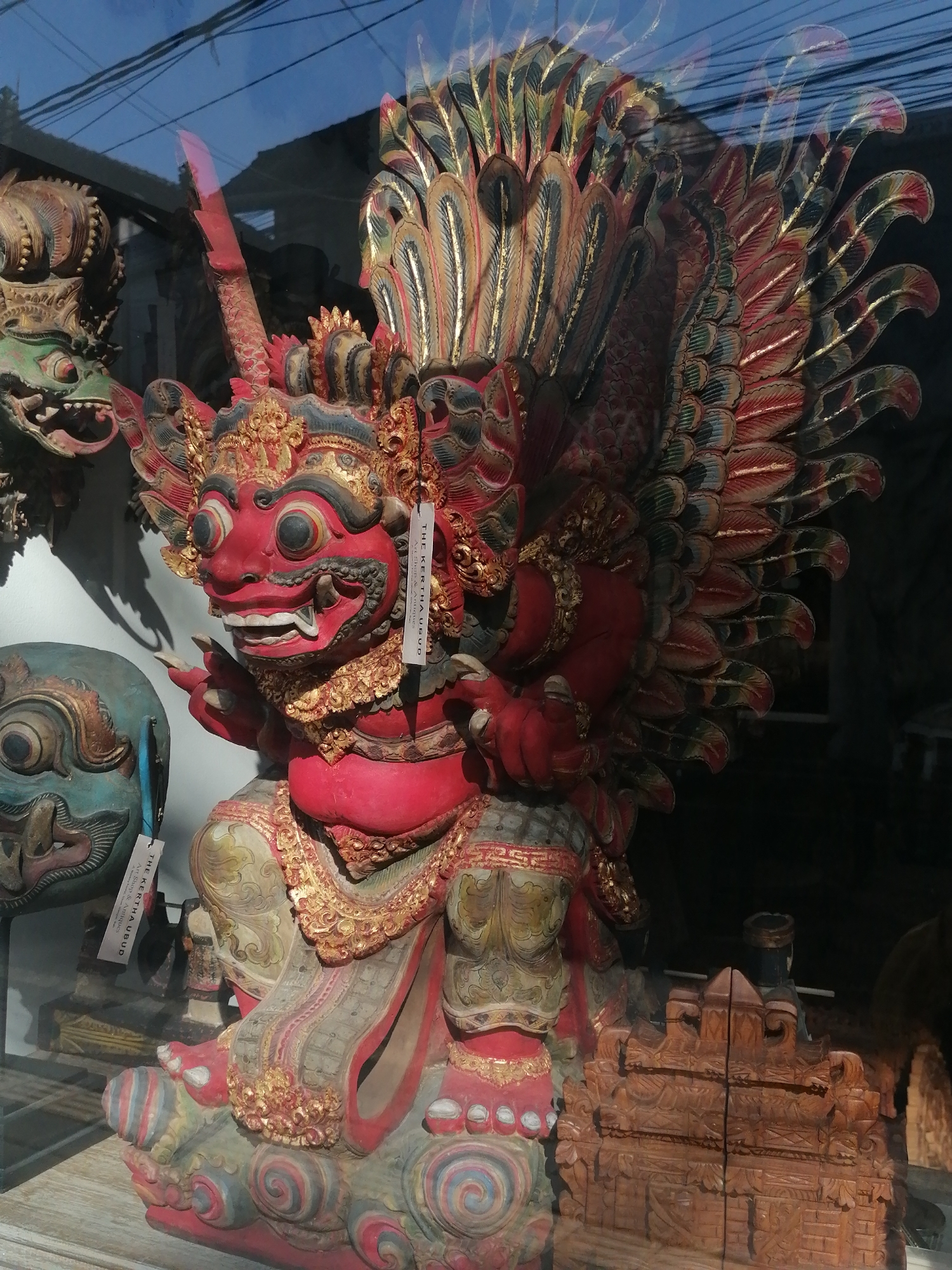 This is a medium shot, taken indoors, of a large, intricately carved wooden statue of a mythological creature, likely a demon or deity, displayed in a shop window in Ubud District, Indonesia. The statue is predominantly red, with accents of gold, green, and blue. It features a fierce facial expression with wide eyes, sharp teeth, and a gaping mouth. Large, feathered wings fan out behind its head, elaborately decorated with red, gold, and green paint. The creature is posed in a seated or kneeling position, with its hands outstretched. The statue is positioned in front of other carved wooden items and masks, including a green-faced mask to the left. A white tag hangs from the main statue, with the text "THE KERTHA Ubud Art Shop & Antiques". The scene is viewed through a glass window, reflecting the sky and overhead wires, indicating it is likely displayed at street level. The lighting is natural, coming from outside.
