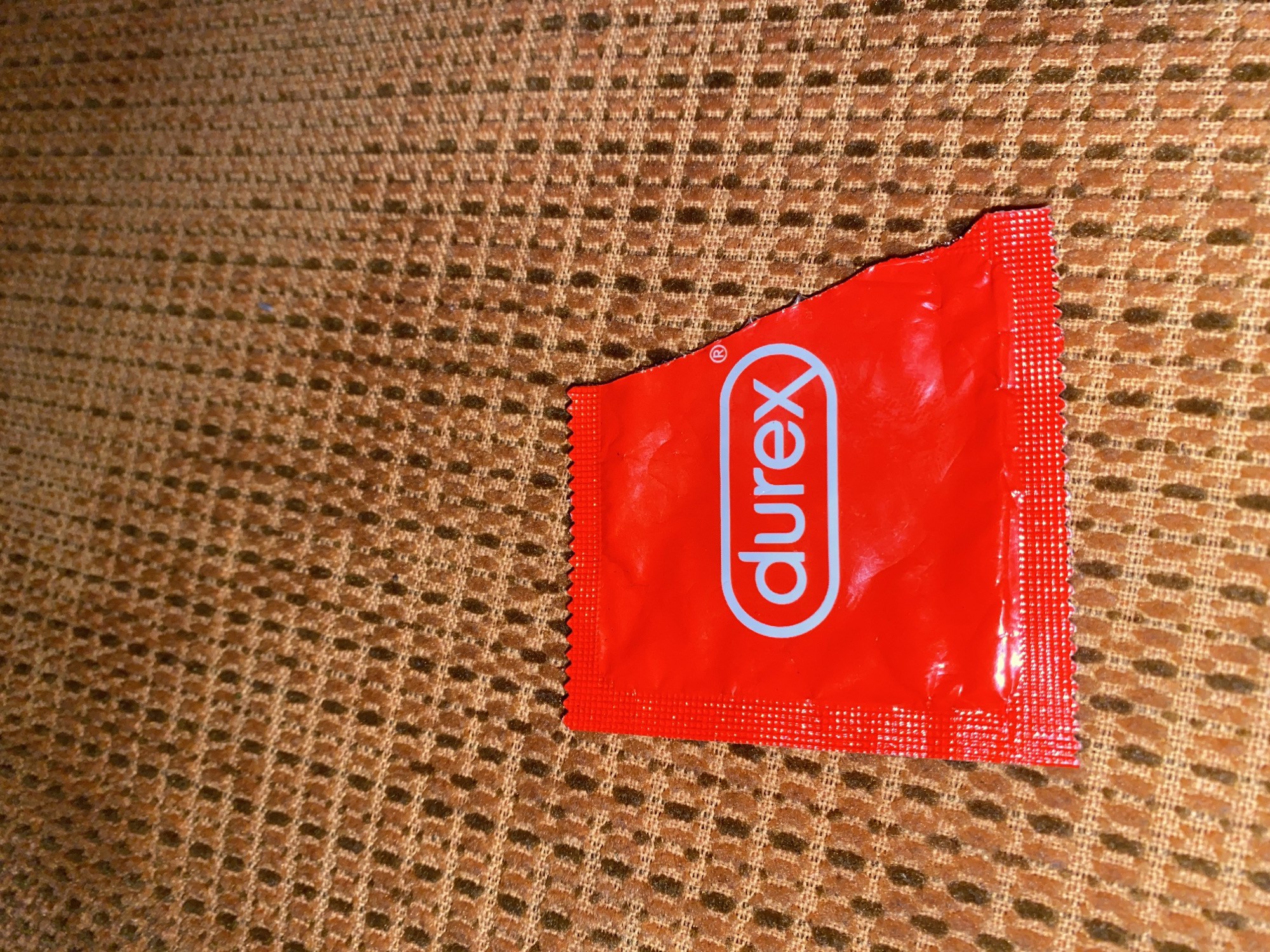 The image displays a torn red condom wrapper with the Durex logo printed in blue. The wrapper is lying on a brown, textured surface, likely a fabric with a woven pattern. The lighting is dim, creating shadows and highlighting the texture of the surface. There are no people or other objects visible in the image. The location in Brashlen, Bulgaria, is not depicted or suggested by any elements in the photograph.