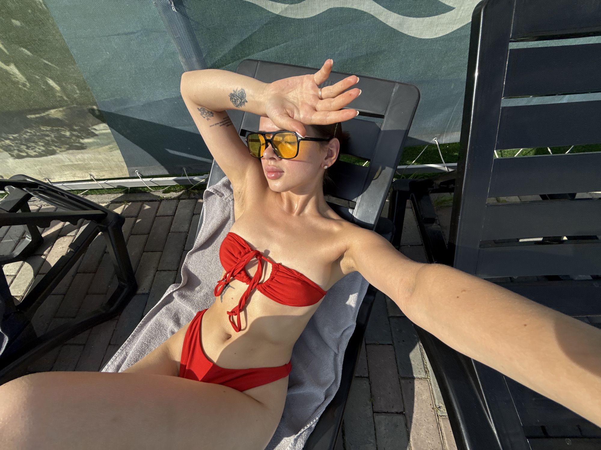 A young woman is lying on a black plastic deck chair outdoors. She is wearing a red bikini and yellow sunglasses. Her right arm is outstretched, taking a selfie, and her left arm is raised to shield her eyes from the sun. A towel is draped over the chair beneath her. Another similar deck chair is visible to the left. The ground is paved with bricks. Overhead, there's a shade structure, likely some type of awning. Greenery, possibly trees or bushes, can be seen behind the shade structure. It is daytime. I do not have enough information to place the exact location within Гамово, Russia.