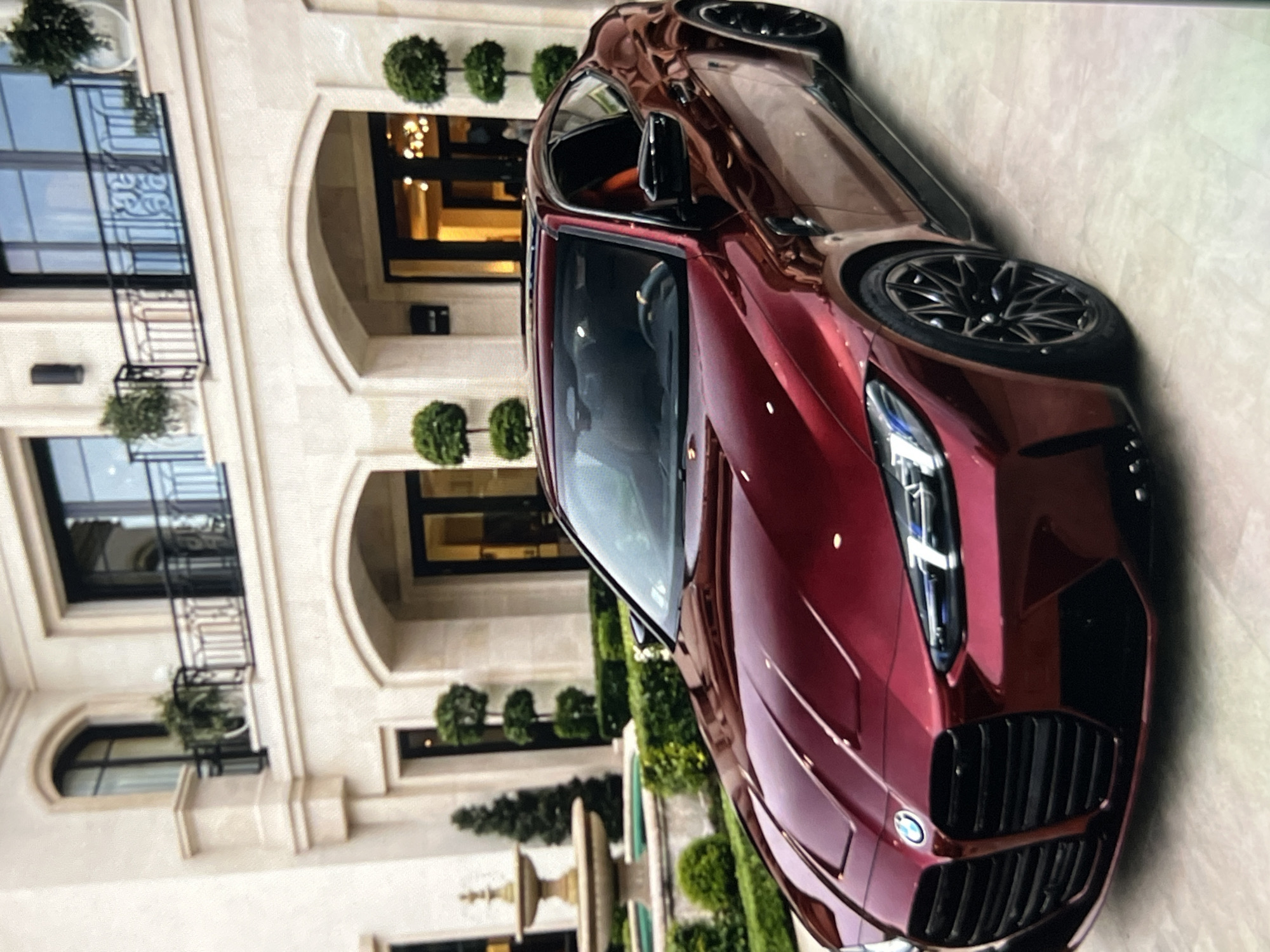 A red BMW M4 is parked on a paved surface in front of a large, modern, light-colored mansion. The mansion has multiple arched doorways, large windows, and balconies with black railings. There is a fountain to the left of the car and manicured bushes and trees surrounding the property. The car is the primary subject, with its hood, grille, headlights, and wheels clearly visible. The sky is not visible in the image.