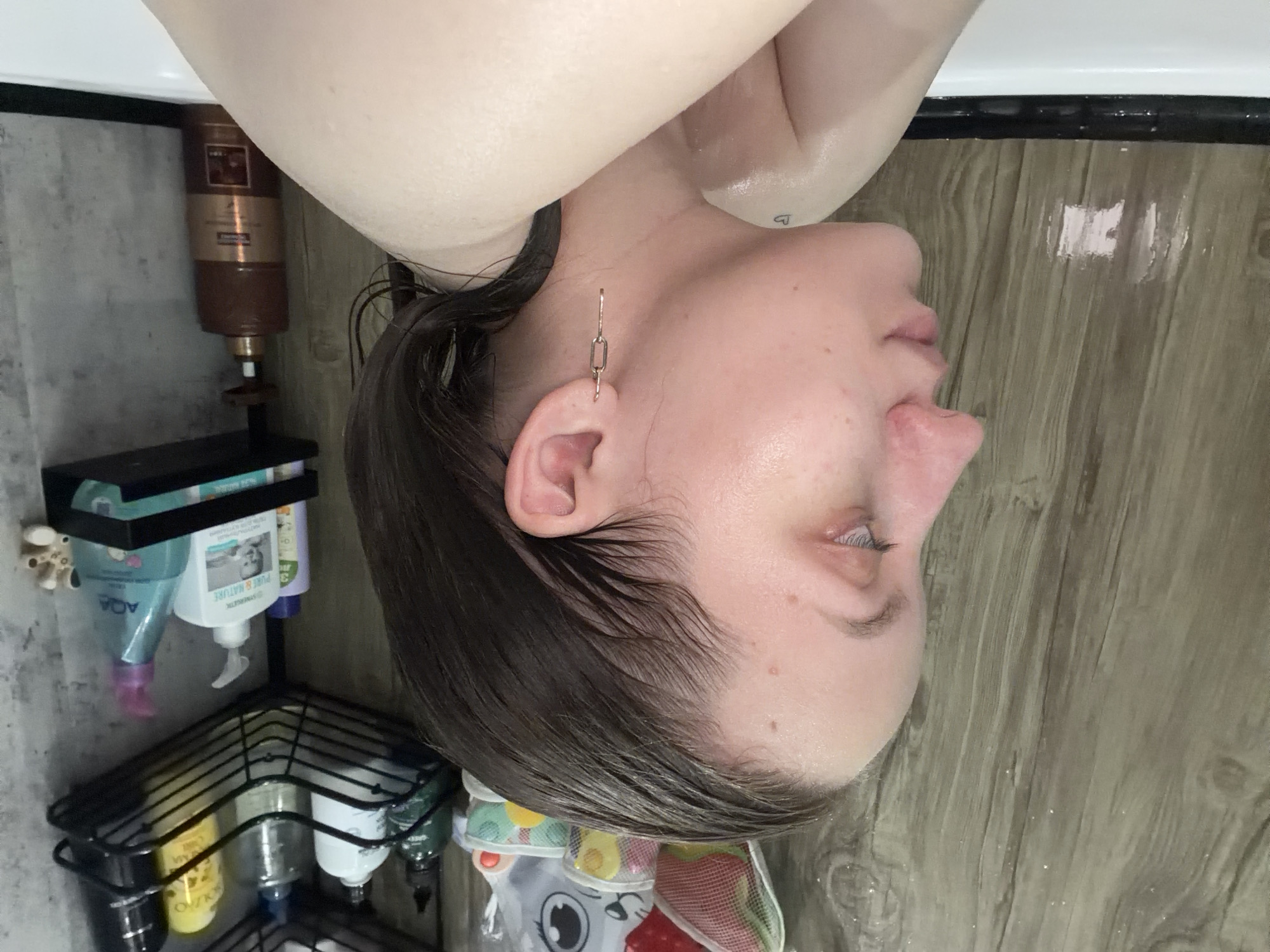 A close-up, rotated image of a woman with wet, dark hair and fair skin is centered in the frame. She is wearing a gold, geometric earring in her left ear. The background includes a partially visible white tub edge at the top, a wood-grain patterned floor on the right, a gray wall to the left, and a metal shelving unit attached to the wall containing various toiletry bottles. A brown pump bottle sits above the shelving unit. The image suggests the woman is in a bathroom. There is no indication of the location being Reshetnikovo, Russia.