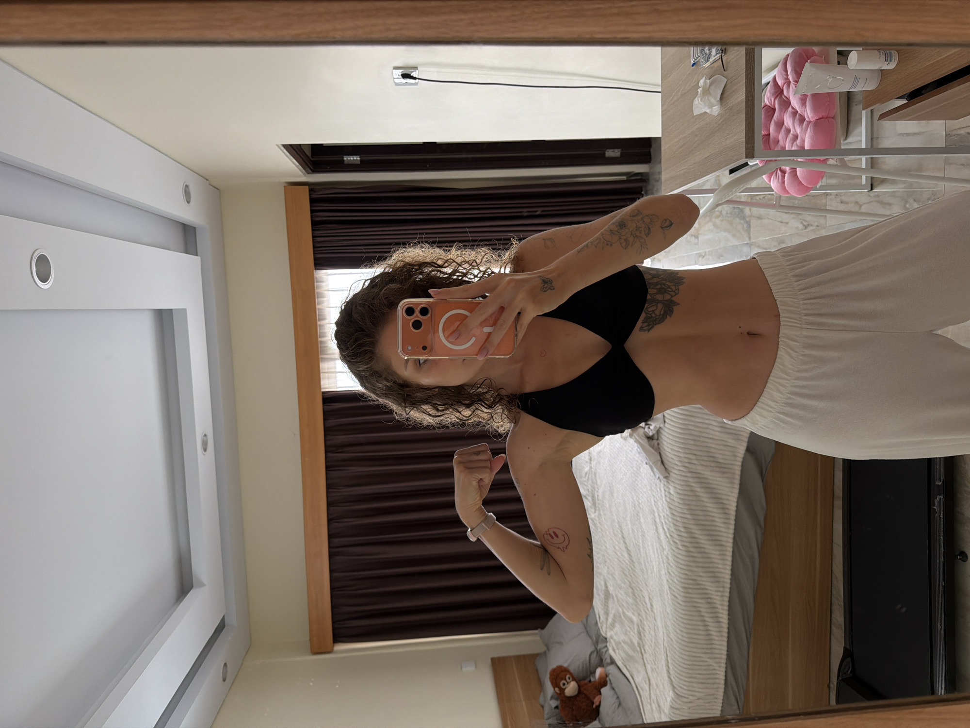 The image shows a woman with curly brown hair, wearing a black bikini top and white loose-fitting pants. She is standing in what appears to be a bedroom or hotel room, taking a mirror selfie with her phone. The room has a bed with striped bedding, a dresser, a treadmill, and dark brown curtains. A stuffed monkey toy is visible on the bed. In the reflection, a table with some items on it, including a pink cushion, is also visible. The overall impression is that of a casual, domestic setting. The location is specified as North Kuta, Indonesia, suggesting this is likely a hotel room or apartment in that area.