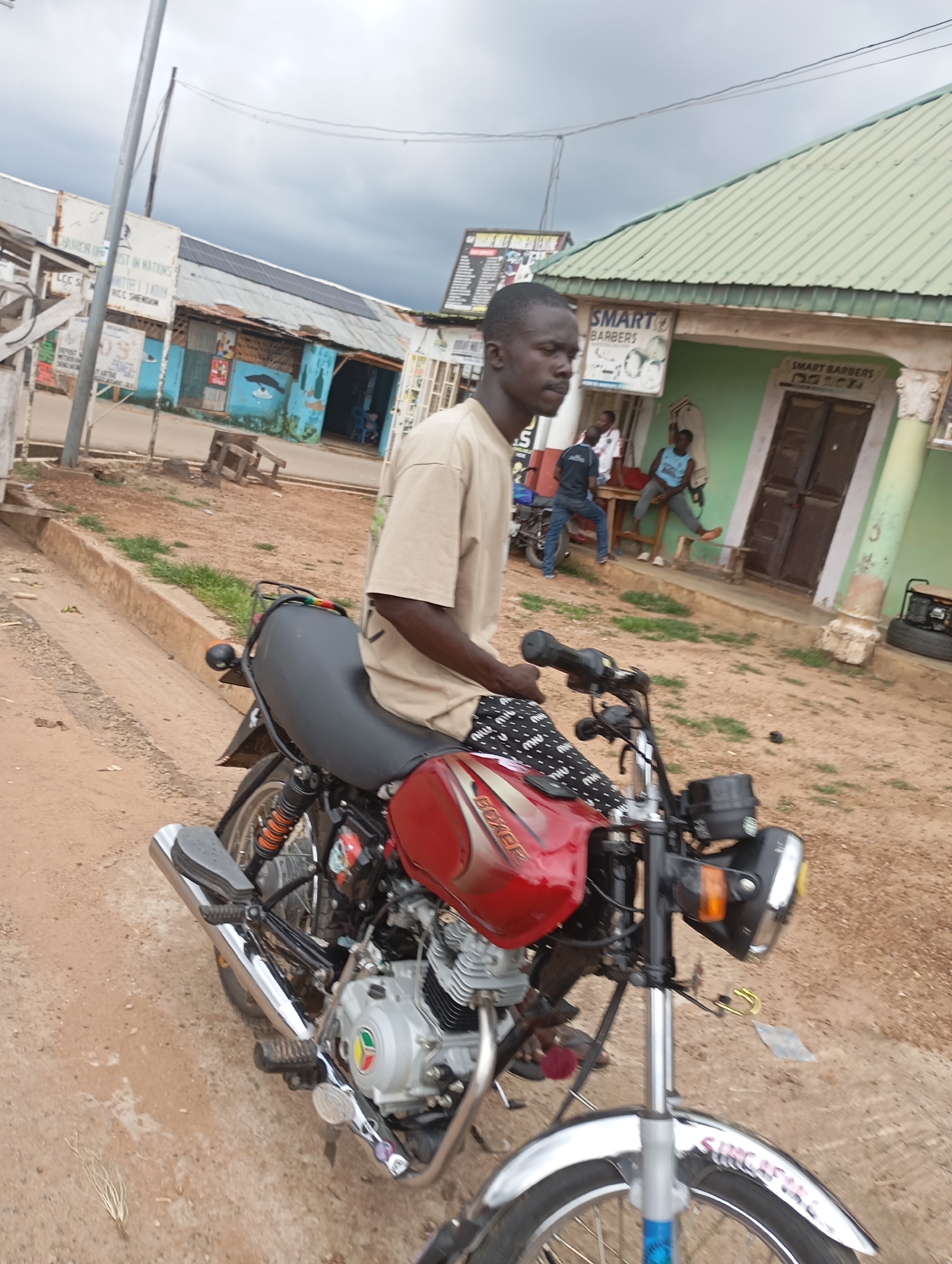 In Shendam, Nigeria, a young Black man wearing a tan t-shirt and patterned shorts is seated on a red and black motorcycle. The motorcycle's engine is visible, and it has a chrome exhaust pipe. The man is looking to his right. In the background, several other people are visible, some standing and some sitting, near a building with a green roof that has "SMART BARBERS" written on it. Another building with blue and white siding is also visible. The ground is dirt and sand. The sky is overcast.