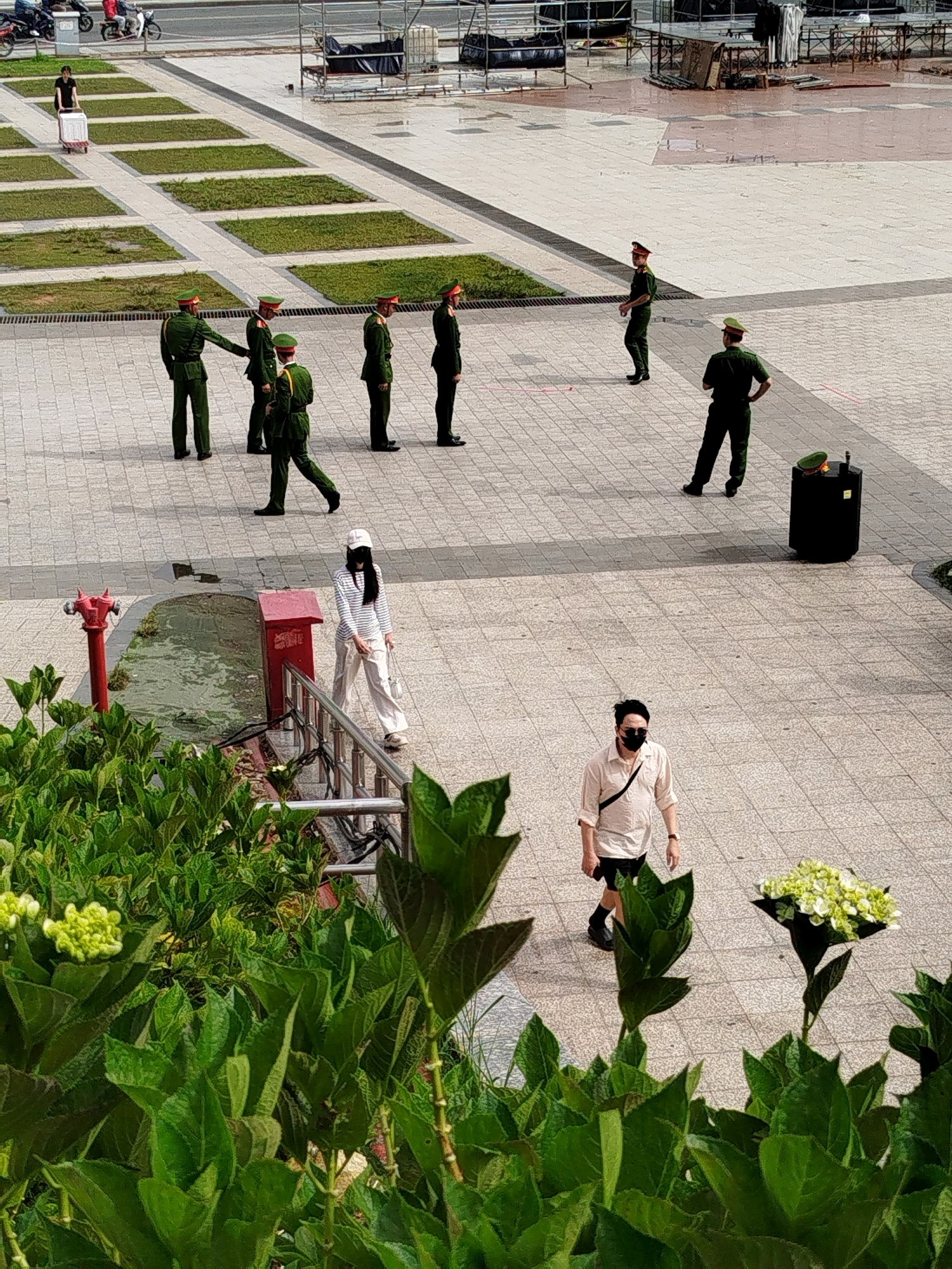 In Da Lat, Vietnam, a group of individuals in green uniforms, likely police officers, are standing on a paved plaza. Some are in formation, while others are dispersed. A person in a white and black striped shirt and white pants, wearing a white cap, is walking past a red fire hydrant and a red mailbox. Another person wearing a light-colored shirt, black shorts, black socks, and a black face mask is walking across the plaza. In the foreground, lush green foliage, possibly hydrangeas, obscures the lower part of the scene. The plaza appears to be a public space with distinct square garden beds and a wet, reflective surface in the background, suggesting recent rain or cleaning.