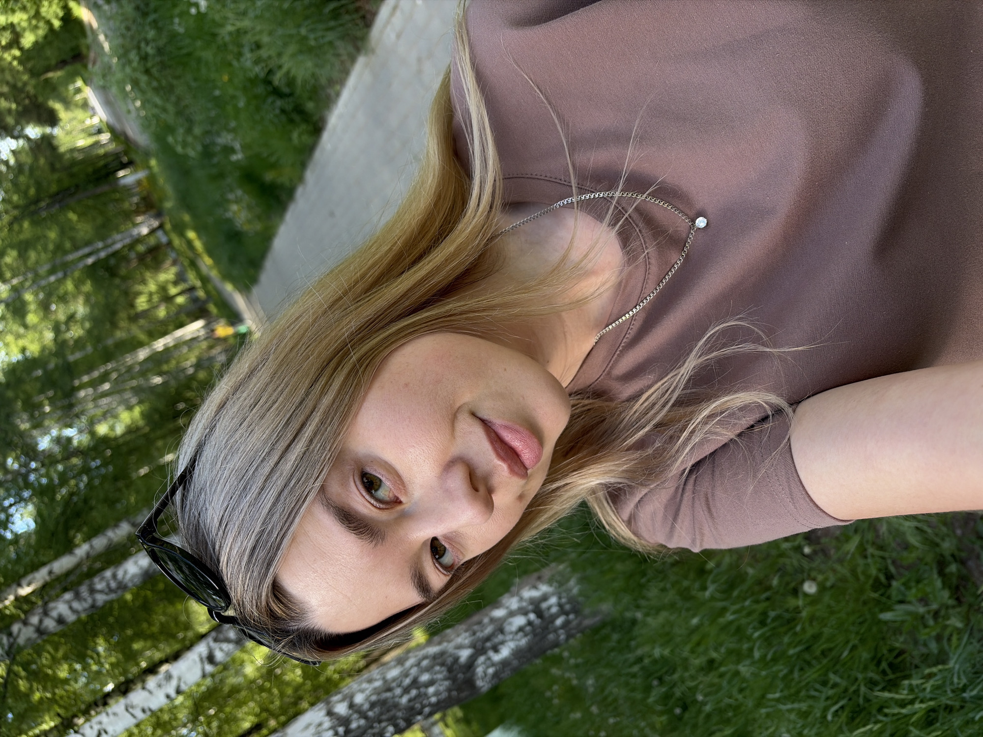 A woman is pictured outdoors. She has light blonde hair with darker roots. She's wearing a brown top and a silver necklace with a small pendant. Her sunglasses are perched on her head. She is facing the camera with a slight smile. The background consists of trees and greenery, suggesting a park-like setting. The image was taken in Chaikovsky, Russia.