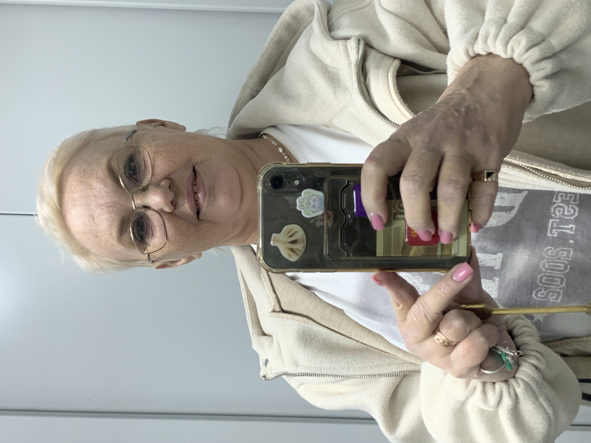 A woman with short, light blonde hair and glasses is taking a selfie. She is wearing a white t-shirt, a beige hooded jacket, and several rings. Her fingernails are painted pink. The phone she is using has a sticker on the back resembling a dumpling. The text on her t-shirt is partially obscured but appears to read "SOOS T". The location and city of Chaikovsky, Russia cannot be determined from the provided image.