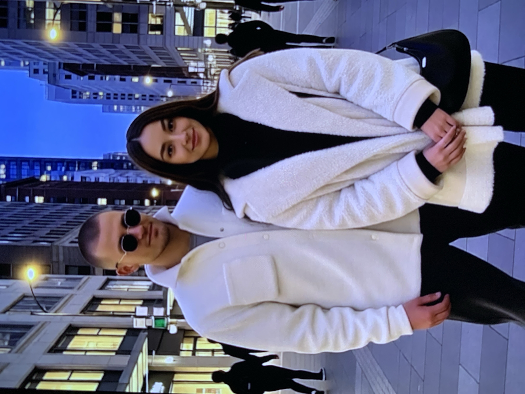A young Caucasian couple is standing on a city sidewalk at dusk. The man, on the left, is wearing a white jacket and sunglasses. He has short brown hair and is looking to the left. The woman, on the right, is wearing a white coat and has long brown hair. She is looking at the camera and smiling. The background is a city street with tall buildings and streetlights. The sky is a deep blue, indicating that it is either dawn or dusk. The sidewalk is paved with grey tiles. There are other people in the background, but they are out of focus. The image is well-lit, and the subjects are in focus.