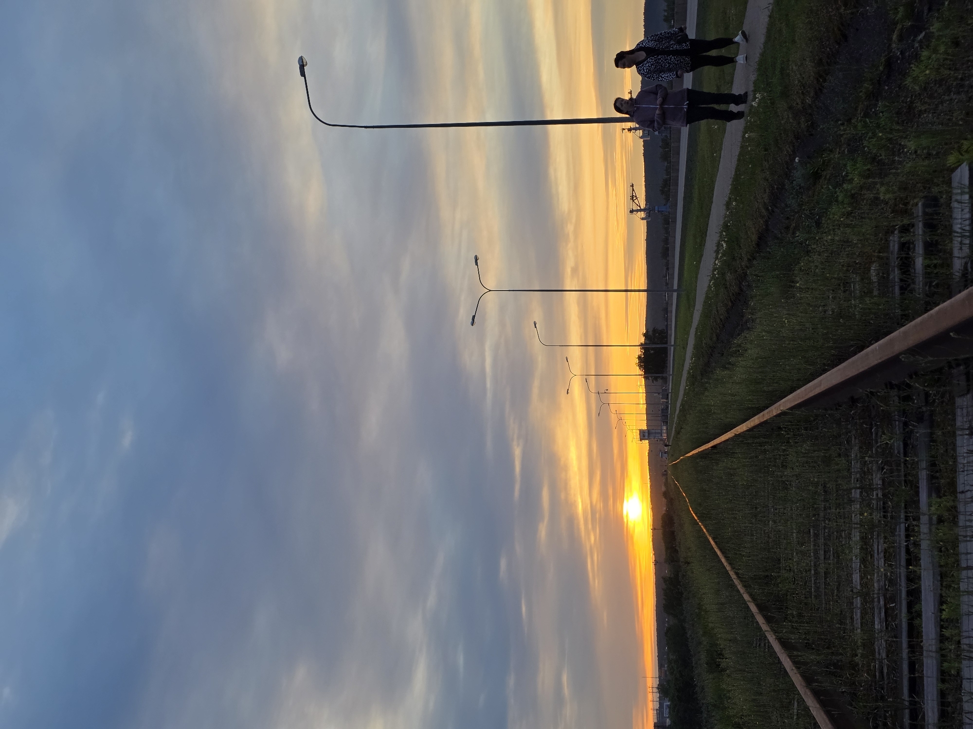 The photograph, taken in Chaikovsky, Russia, presents an elongated view of a sunset. The sky is a blend of blue and orange hues. The setting sun is positioned at the horizon, its light casting a bright glow. Along the right side of the image, figures of two people are visible, walking along a path next to a roadside. A series of streetlights punctuate the horizon. In the foreground, a metallic fence-like structure runs along the perspective lines, with vegetation growing on either side.