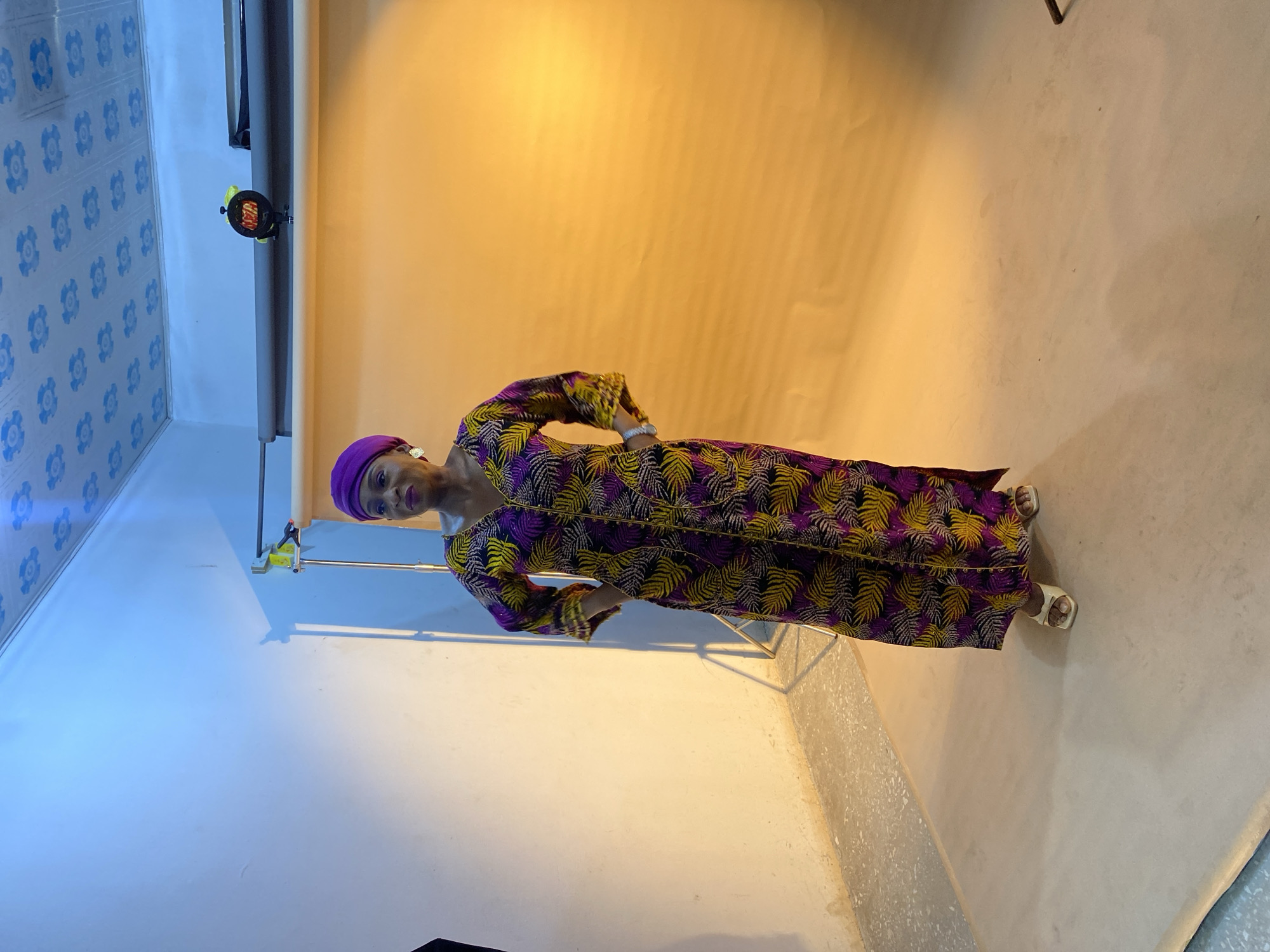 A woman stands in a room with a textured blue and white tile ceiling. She wears a purple headscarf, a long dress patterned with yellow and purple leaf designs, and white sandals. The background is a combination of a peach-colored wall and a white wall. Photography equipment, including a light stand and reflector, is visible. The location is Gura Top, Nigeria.
