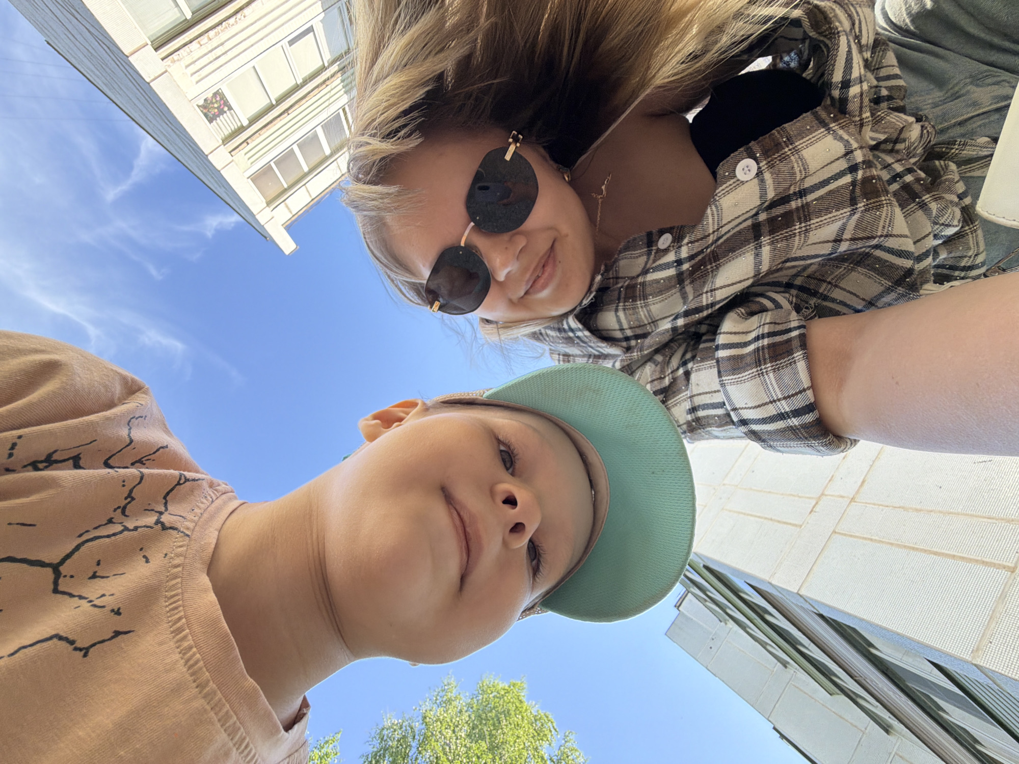 Two people, a young woman and a child, are looking down at the camera. The woman is wearing sunglasses and a plaid shirt. The child is wearing a light green cap and a pink shirt. The background includes a blue sky, buildings, and trees. The location is likely an urban area in Chaikovsky, Russia, based on the architectural style of the buildings.