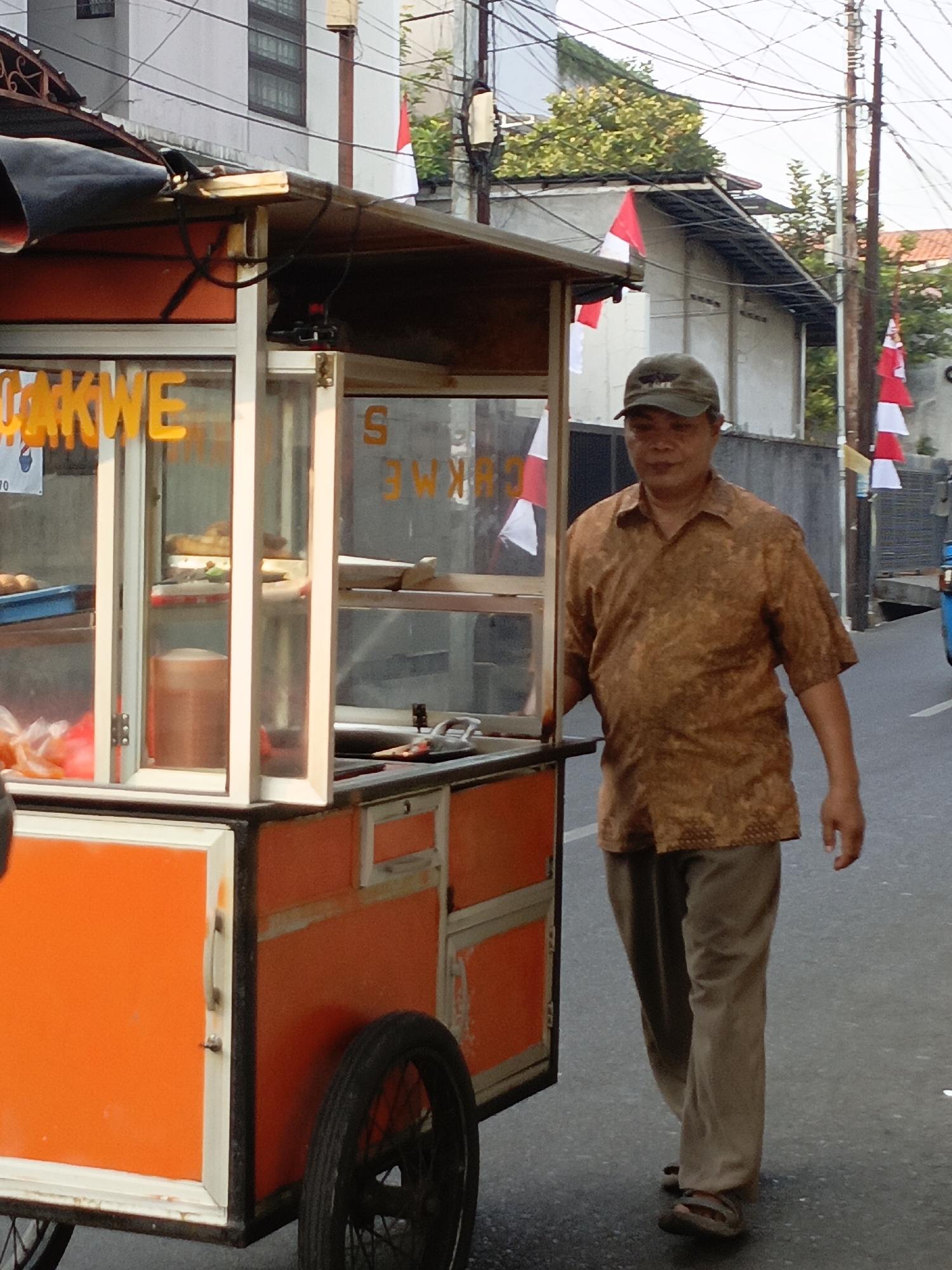 A man in a brown patterned shirt and tan pants stands next to an orange food cart. The food cart has glass display cases and is parked on a street. The man is wearing a dark baseball cap and sandals. In the background, there are buildings, trees, and Indonesian flags. The man appears to be walking away from the cart. The scene takes place in the city of East Jakarta, Indonesia.