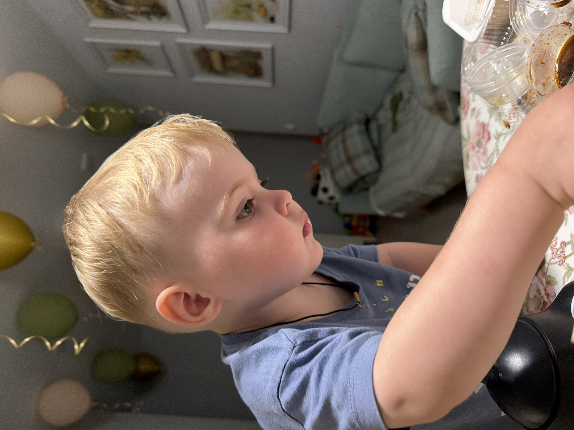 The media file is a close-up, angled photograph of a young Caucasian child with blonde hair. The child is wearing a blue t-shirt and appears to be looking off-camera to the right. The child's left ear and a portion of their arm are visible. In the background, there are several balloons, some pale pink and some green, with gold ribbon streamers. The setting appears to be indoors, possibly a home or a party venue, with framed pictures on the wall and a floral-patterned tablecloth visible on a surface to the right. A dark object, possibly a speaker or toy, is also partially visible in the foreground on the right. The lighting suggests it could be daytime. The child's expression is neutral, and the overall mood seems relaxed. There is no visible text in the image. The location context is Votkinsk, Russia.