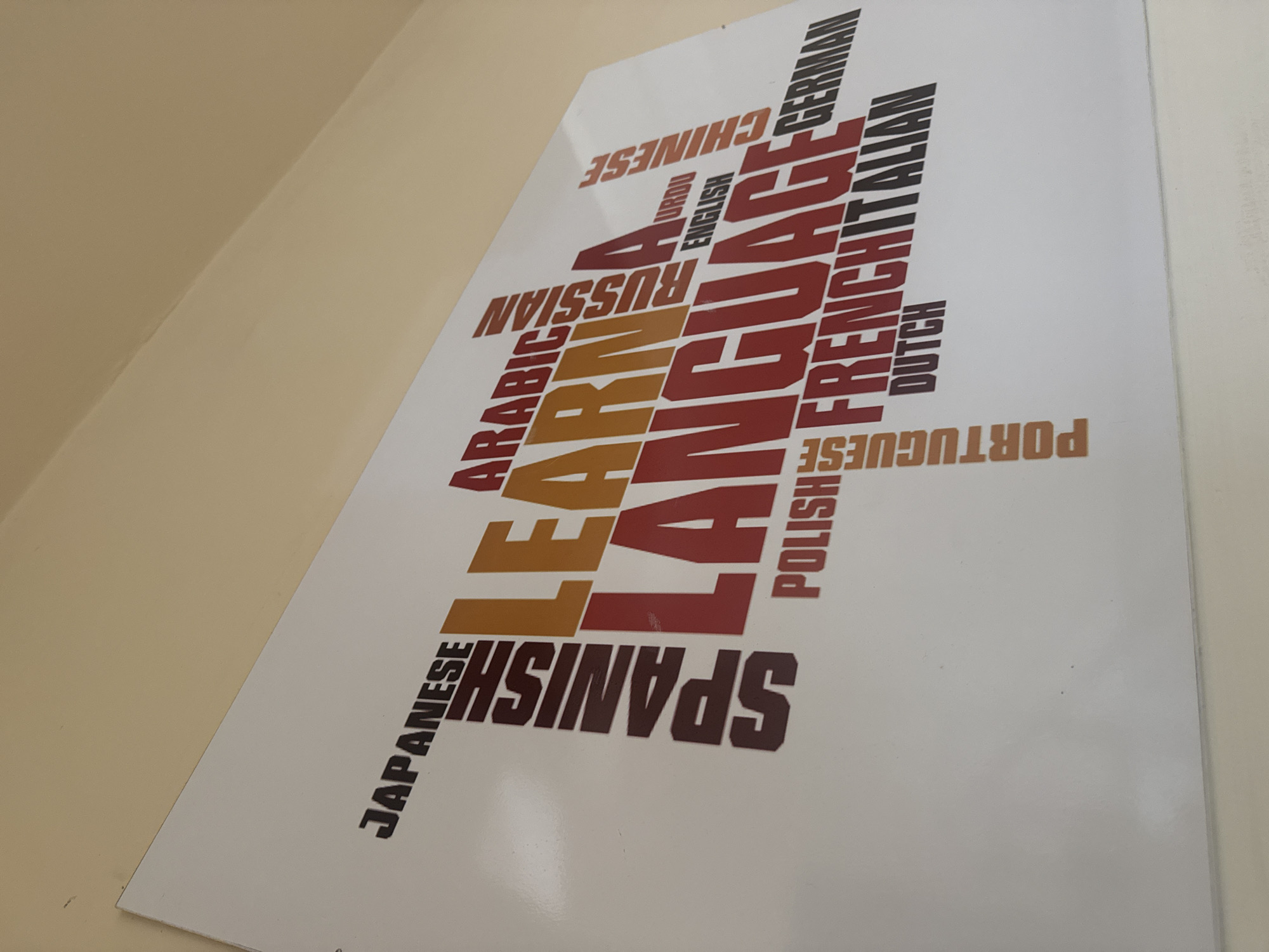 The image displays a white rectangular sign or poster affixed to an off-white or beige wall. The sign features a word cloud design composed of various language names and the phrase "LEARN A LANGUAGE." The words "LEARN" and "LANGUAGE" are prominently displayed in large, bold, sans-serif fonts, with "LEARN" in orange-brown and "LANGUAGE" in red. Numerous other language names are interspersed around these central words, varying in size, orientation, and color (dark brown, black, orange-brown, red). Identifiable language names include Japanese, Spanish, Arabic, Russian, Urdu, English, Chinese, German, Polish, French, Italian, Portuguese, and Dutch. The sign is angled slightly, suggesting it is mounted high on the wall, with the upper right corner receding into the background. The wall surface appears smooth and clean, with a subtle texture visible. No people or interactions are present in the scene.
Location: Unknown, Unknown.