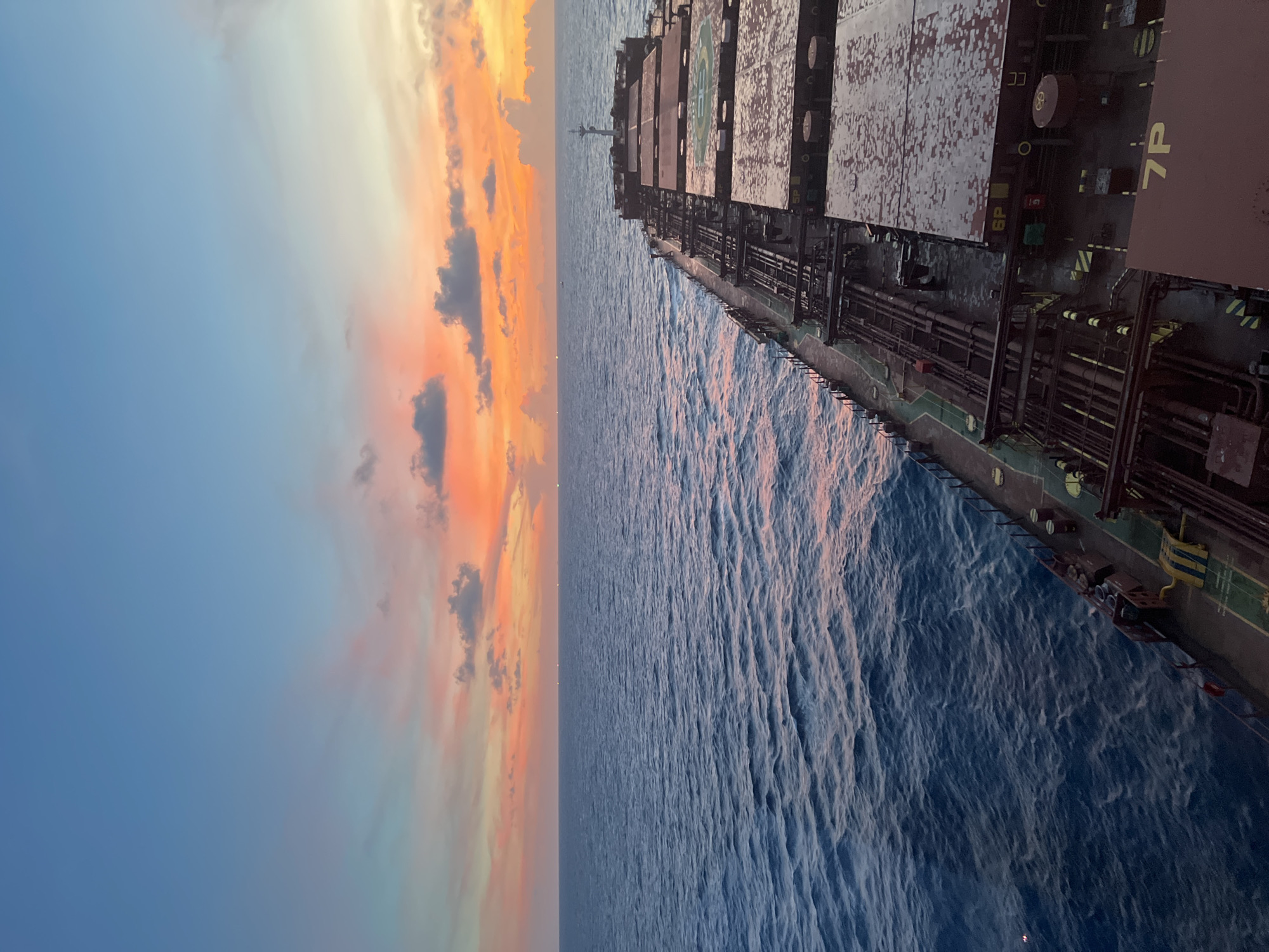 The image shows a view from the deck of a large cargo ship at sea during sunset. The sky is a gradient of blue, pink, and orange, with scattered clouds. The sea is calm with gentle waves. The ship's deck is visible in the foreground, with its cargo holds and piping. There are no people visible in the image. The scene is set in an unknown location at sea.