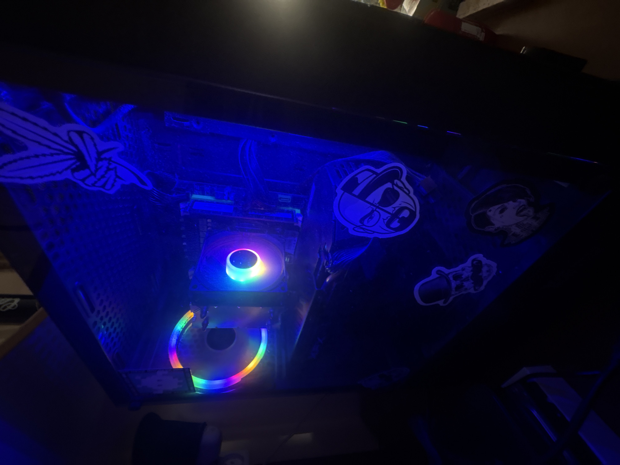 The image shows the inside of a computer tower case, illuminated by blue and rainbow-colored LED lights from the fans and other components. The case has a clear side panel, allowing the internal components to be visible. Several stickers are affixed to the inside of the case. One sticker, near the top, appears to be a stylized image of two fingers forming a peace sign. Below this, another sticker depicts a character with a hat and eye patch. Further down, a sticker of a skull wearing a top hat and mustache is visible, and near the bottom, a sticker shows a profile of a person with dark hair. The computer components are dark and black, with the lights creating a vibrant, futuristic aesthetic. There are no people visible in the image. The scene is entirely indoors, with no visible connection to Sverdlovskiy, Russia.