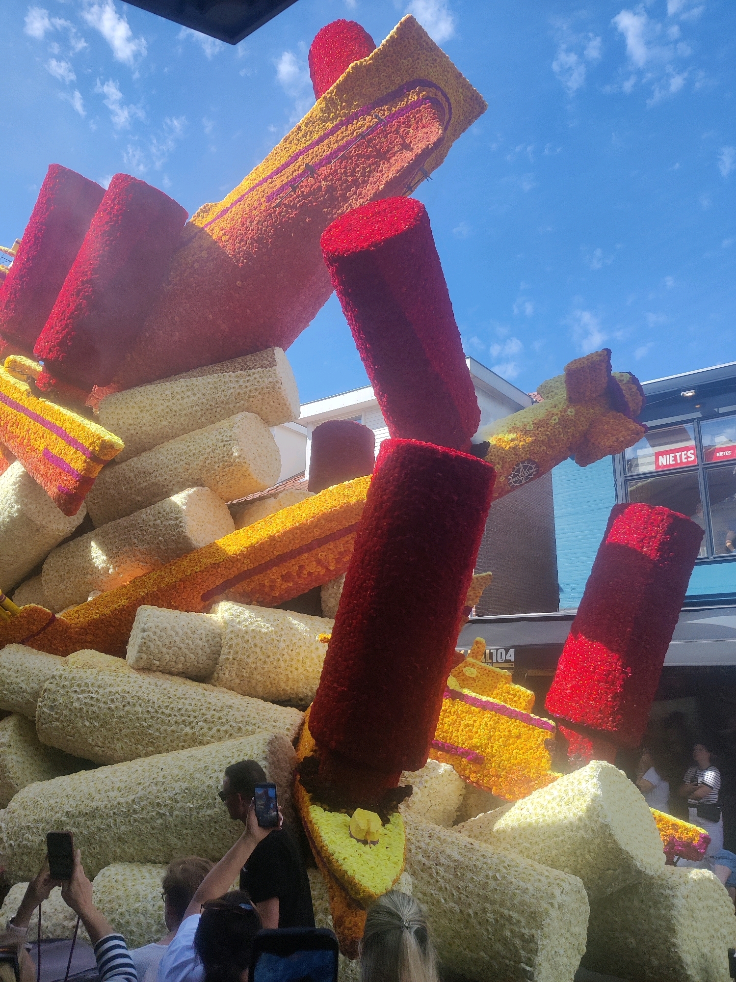 An outdoor, eye-level, slightly upward-angled medium shot captures a large, colorful floral float constructed from many different types of flowers, including red, yellow, and white varieties. The float is intricately designed, featuring cylinder-like shapes and larger rectangular structures, all meticulously covered in the flowers. In the background, buildings with windows and a blue sky with scattered white clouds are visible. Some of these buildings have signage. The foreground shows the heads and shoulders of several people watching the float, some of whom are holding up their phones to take pictures. One person in the middle foreground is wearing sunglasses and holding a smartphone. The overall scene suggests a parade or festival event taking place in Zundert, Netherlands, as indicated by the presence of the float.