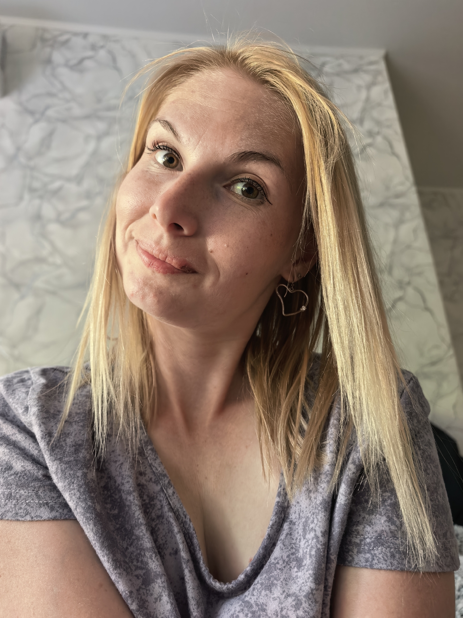 A fair-skinned woman with blonde hair is the subject of this close-up shot. She has green eyes and some freckles visible on her face. She is wearing heart-shaped earrings and a patterned gray T-shirt with a V-neck. The background is a marble-like textured wall. There is no evidence that this photo was taken in Sterlitamak, Russia.