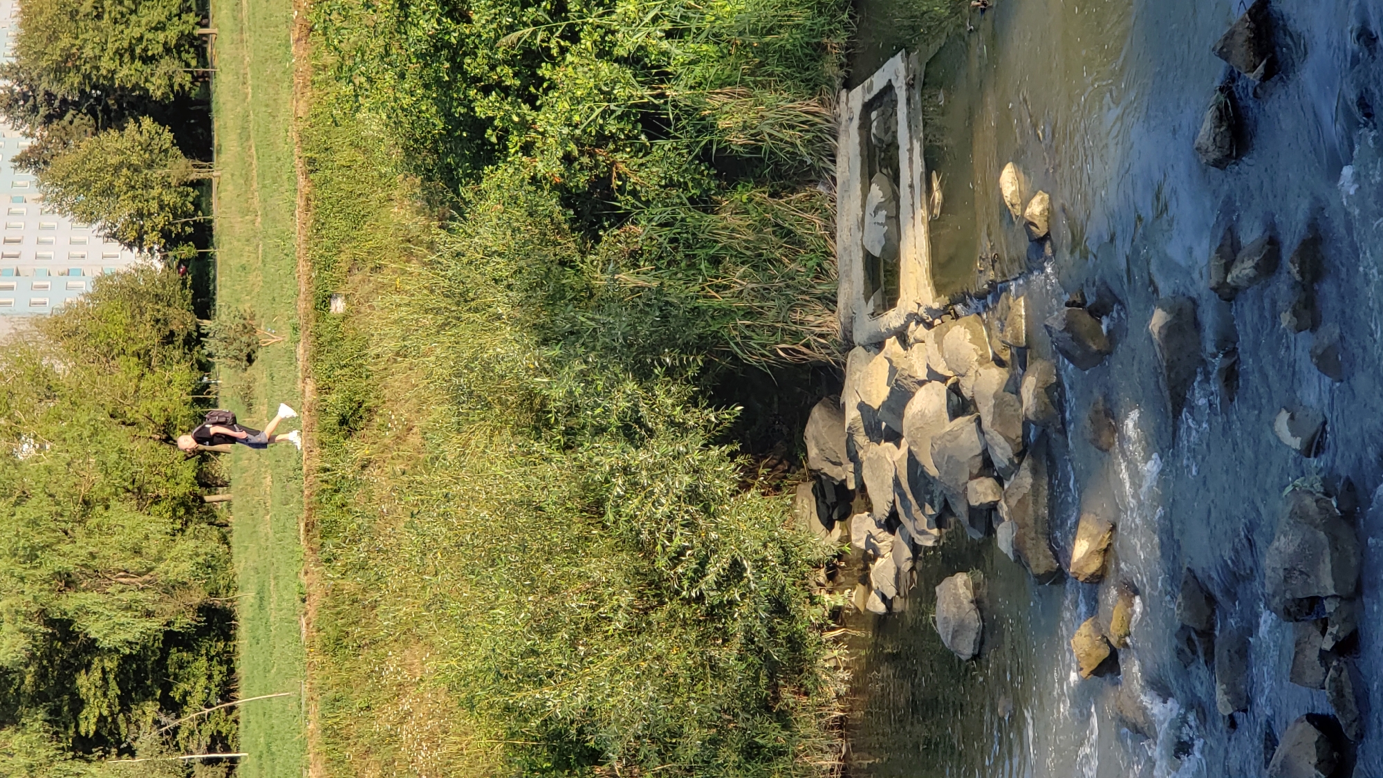 A man walks on a path beside a grassy embankment. He is wearing a black t-shirt, blue shorts, white sneakers, and a backpack. The path is lined with grass and trees. In the foreground, a shallow river flows over rocks. There is a concrete structure with water in it to the right of the river. In the background, a building with light-colored walls and many windows is visible. The image was taken in the area of Sibírska street in Prešov, Slovakia.