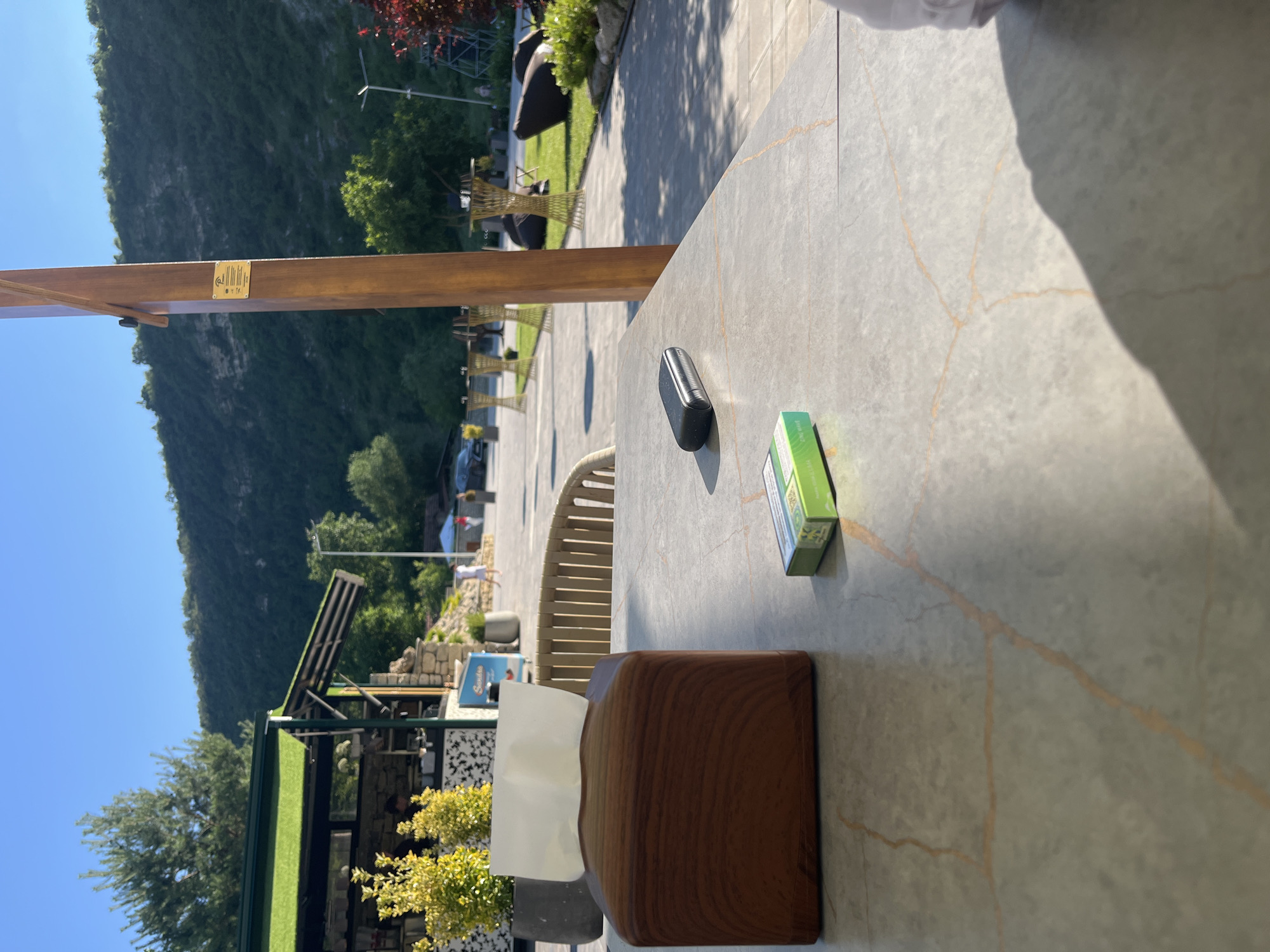 The image depicts an outdoor scene, likely a cafe or restaurant, in Butuceni, Moldova. The orientation of the image is landscape, with the sky and a verdant hillside on the left. In the foreground, a marble-topped table is visible, with a wooden tissue box and a small green box, possibly containing cigarettes or nicotine pouches, placed on it. A dark, elongated object, which could be an e-cigarette or a pen, also rests on the table. The background reveals a paved area with outdoor furniture and glimpses of greenery and a building with a green roof, suggesting a commercial establishment. The bright sunlight and clear blue sky indicate it is daytime. The overall atmosphere appears relaxed and pleasant, consistent with an outdoor leisure setting. The hillside in the background suggests a location with natural beauty. There is a gold plaque on a wooden beam in the upper left of the image, but the text on it is too small to be legible.