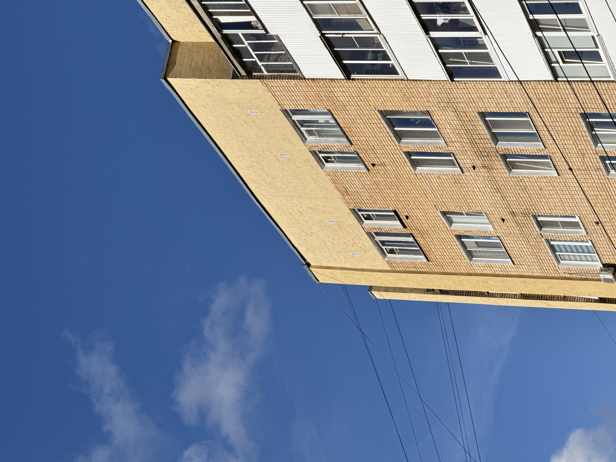 The image shows the exterior of a multi-story apartment building against a bright blue sky with sparse white clouds. The building has sections with yellow stucco and light brown brickwork. There are multiple windows with white frames visible on the brick facade. On the right side of the image, a portion of the building is clad in white vertical siding, also featuring several windows. Overhead, dark power lines crisscross the sky in the upper left portion of the frame. An air conditioning unit is mounted below one of the windows on the brick section. The scene appears to be a detail of an urban residential environment, possibly in Sof'ino, Russia.