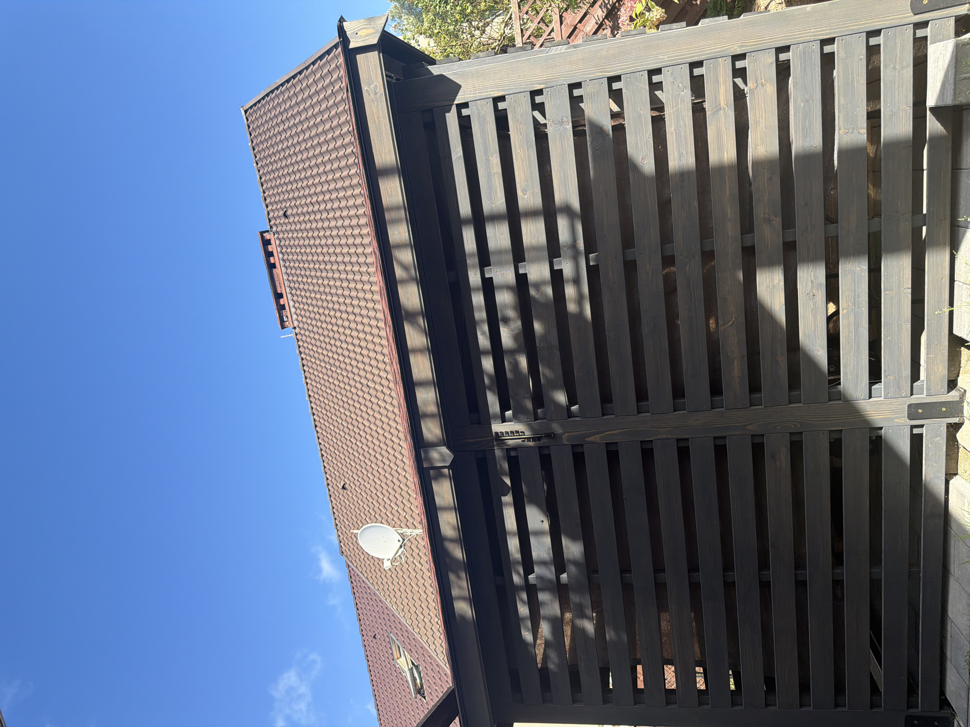 The image shows a view of a dark wooden slatted structure, likely an outdoor storage unit or gazebo, with a tiled roof and a satellite dish visible on the roof. The structure is set against a bright blue sky with a few wispy clouds. The wooden slats are horizontal and spaced apart, allowing glimpses of what is stored inside, possibly firewood. A chimney is also visible on the roof, and what appears to be a skylight. The lighting suggests it is daytime, with shadows cast by the slats and surrounding foliage. No people or specific landmarks are visible that would definitively place the image in Szczecin, Poland.