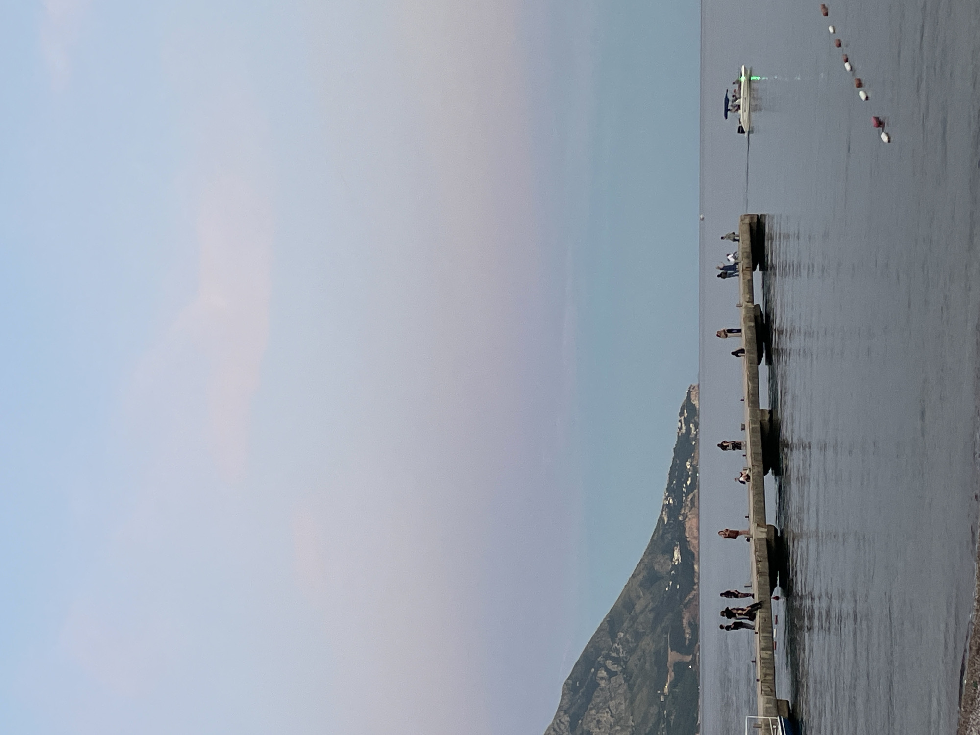 In Budva, Montenegro, a concrete pier extends into calm, dark water under a sky transitioning from pale blue to soft pink and purple hues. Several people are scattered along the pier, some standing and observing the water, others seated or conversing. A small white boat with an awning is moving away from the pier in the distance, leaving a faint green light trail. A mountainous coastline, dotted with buildings, forms the backdrop to the scene. Floating buoys mark a boundary in the water to the right of the pier.