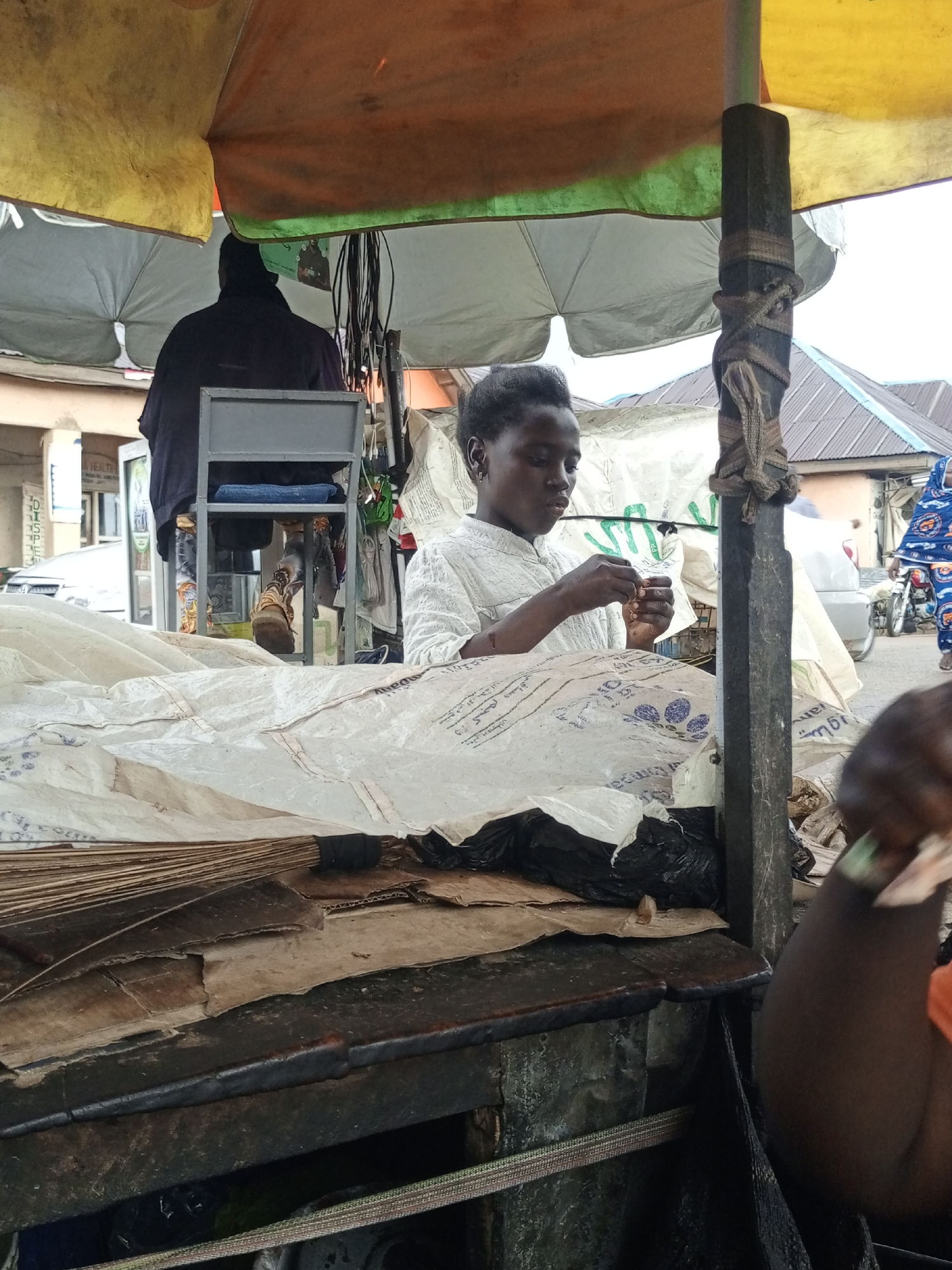 An outdoor market stall is depicted, covered by a yellow and orange tarpaulin. A young Black person, wearing a white top, is seated and engaged in a task with small objects in their hands. Behind them, another person, with their back to the camera, is seated on a chair. The stall displays various items, including what appear to be stacks of paper or cardboard, and plastic sheeting. In the background, buildings with corrugated iron roofs and a motorbike are visible, suggesting an urban setting. The overall impression is that of a typical market scene, possibly in Jos, Nigeria, given the description of the surrounding environment.
