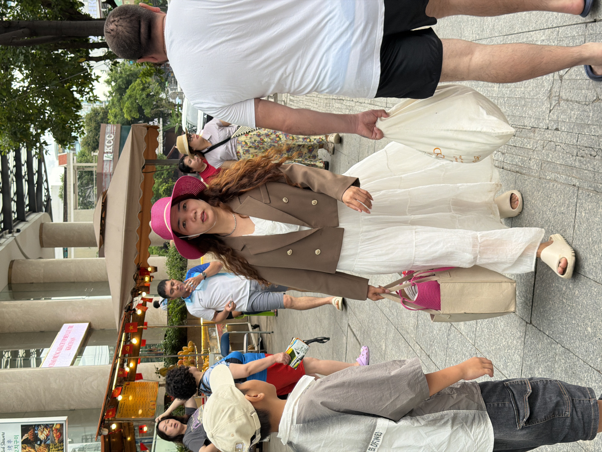 The image shows a street scene in Nha Trang Ward, Vietnam. A woman in a pink hat, brown blazer, and white dress is walking and holding two bags. A man in a white shirt and black shorts is walking behind her, carrying a white plastic bag. Other people, including a man in a blue shirt and a child in a gray shirt, are present in the background. There is a covered outdoor eating area on the left with small Vietnamese flags strung up. The setting is outdoors on a paved area.