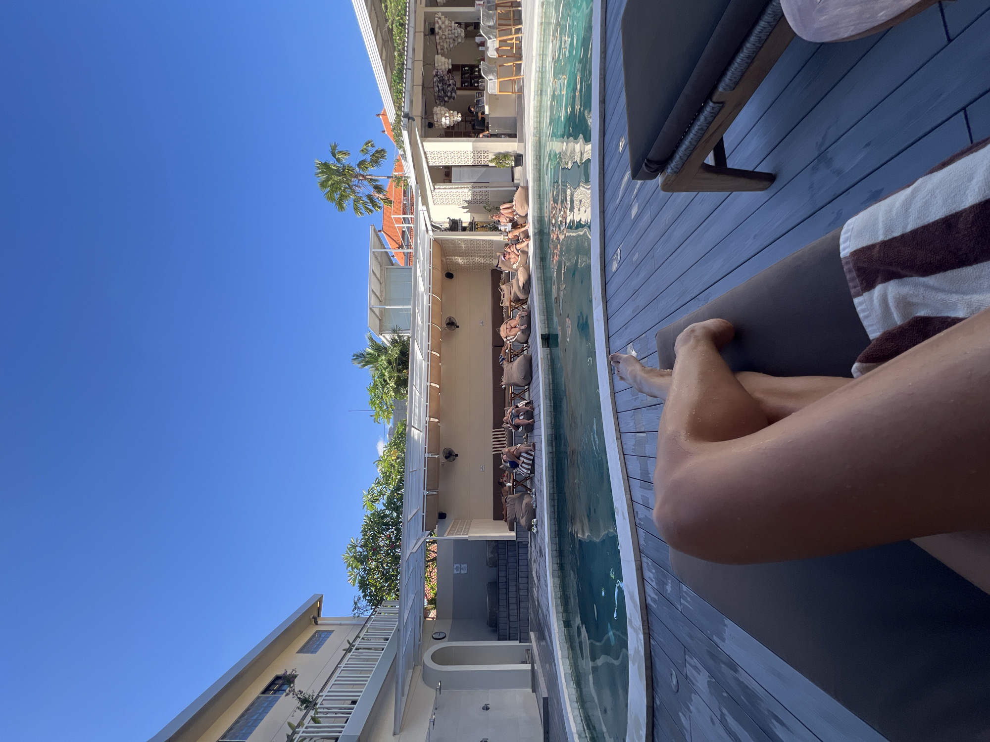 The image shows a daytime scene at a pool-side resort in North Kuta, Indonesia. A partially visible person is relaxing on a lounge chair next to a pool, their legs exposed. The pool runs alongside a building with an open-air lounge area where people are seated in chairs and on cushions. The architecture features white structures with tiled roofs, and tropical trees and palm trees are visible. The sky is clear and blue.