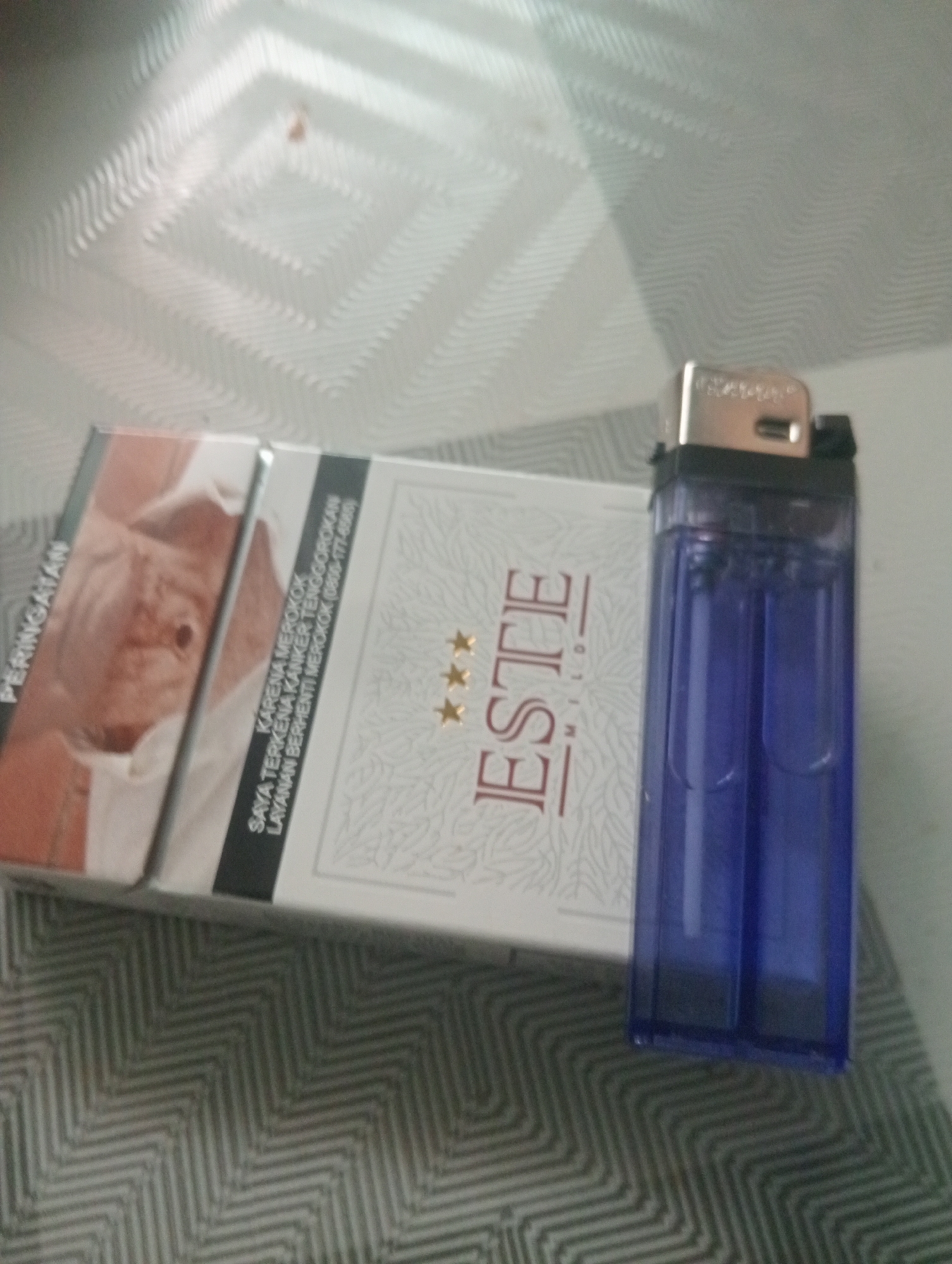 A cigarette pack labeled "ESTE MILD" and a blue disposable lighter are placed on a patterned surface, likely a table or mat. The cigarette pack displays a warning in Indonesian text that translates to "BECAUSE YOU GET CANCER THROAT CANCER GET SMOKING, STOP SMOKING SERVICE (0800-177-6565)". The background consists of a textured, light-colored surface with a diamond pattern. The lighting suggests an indoor environment, possibly a room. The image has a shallow depth of field, with the focus on the cigarette pack and lighter, while the background is blurred. The scene depicts a still life arrangement of smoking paraphernalia.