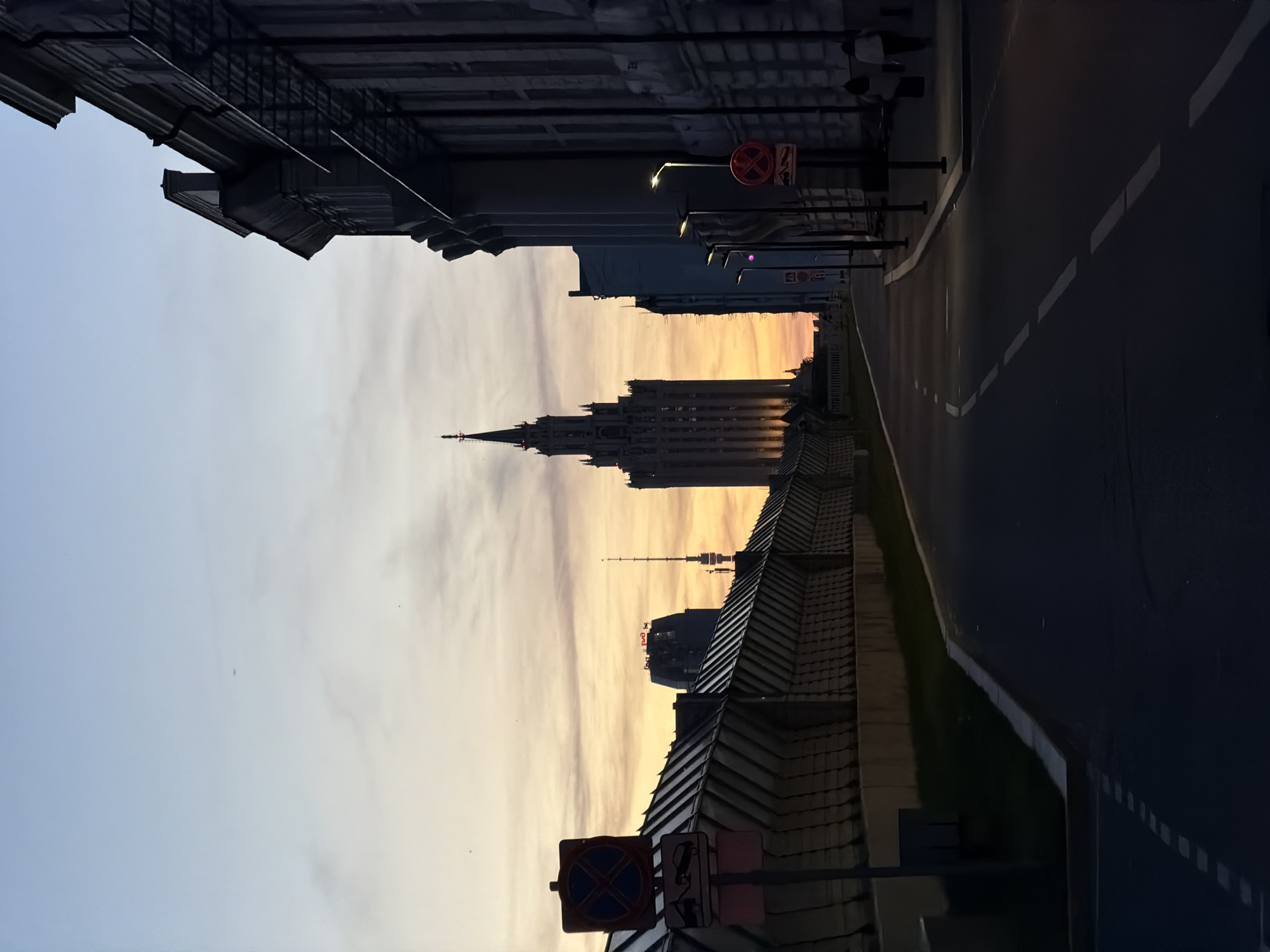 This image depicts an urban street scene in Moscow, Russia, captured at dusk. The sky presents a gradient from light blue at the top to warm orange and yellow hues near the horizon, with scattered faint clouds.
The dominant structure in the mid-ground is the Ministry of Foreign Affairs of Russia building, a tall Stalinist-era skyscraper featuring a distinctive central spire. It is largely silhouetted against the sunset, with some internal lights visible in its lower sections. To the left of this main building, further in the background, a modern high-rise building with a red "РЖД" (Russian Railways) logo on its upper facade is visible, alongside a slender communication tower with an antenna.
The foreground features an asphalt road, curving gently to the right, marked with dashed white lane lines that recede into the distance. Multiple streetlights with illuminated white lamps line the right side of the road, casting localized light pools. On the immediate right, a multi-story building with classical architectural elements, including cornices and downspouts, stands in deep shadow. Its facade appears partially covered or under construction. Two individuals are visible as silhouettes on the sidewalk to the right, walking away from the viewer in the mid-ground.
On the left side of the road, a long, sloped barrier or wall with a corrugated, metallic surface runs parallel to the street. In the left foreground, two vertically stacked traffic signs are present: a "No Stopping" sign (red cross on blue circle) positioned above a "Tow-Away Zone" sign (stylized image of a vehicle being towed).
