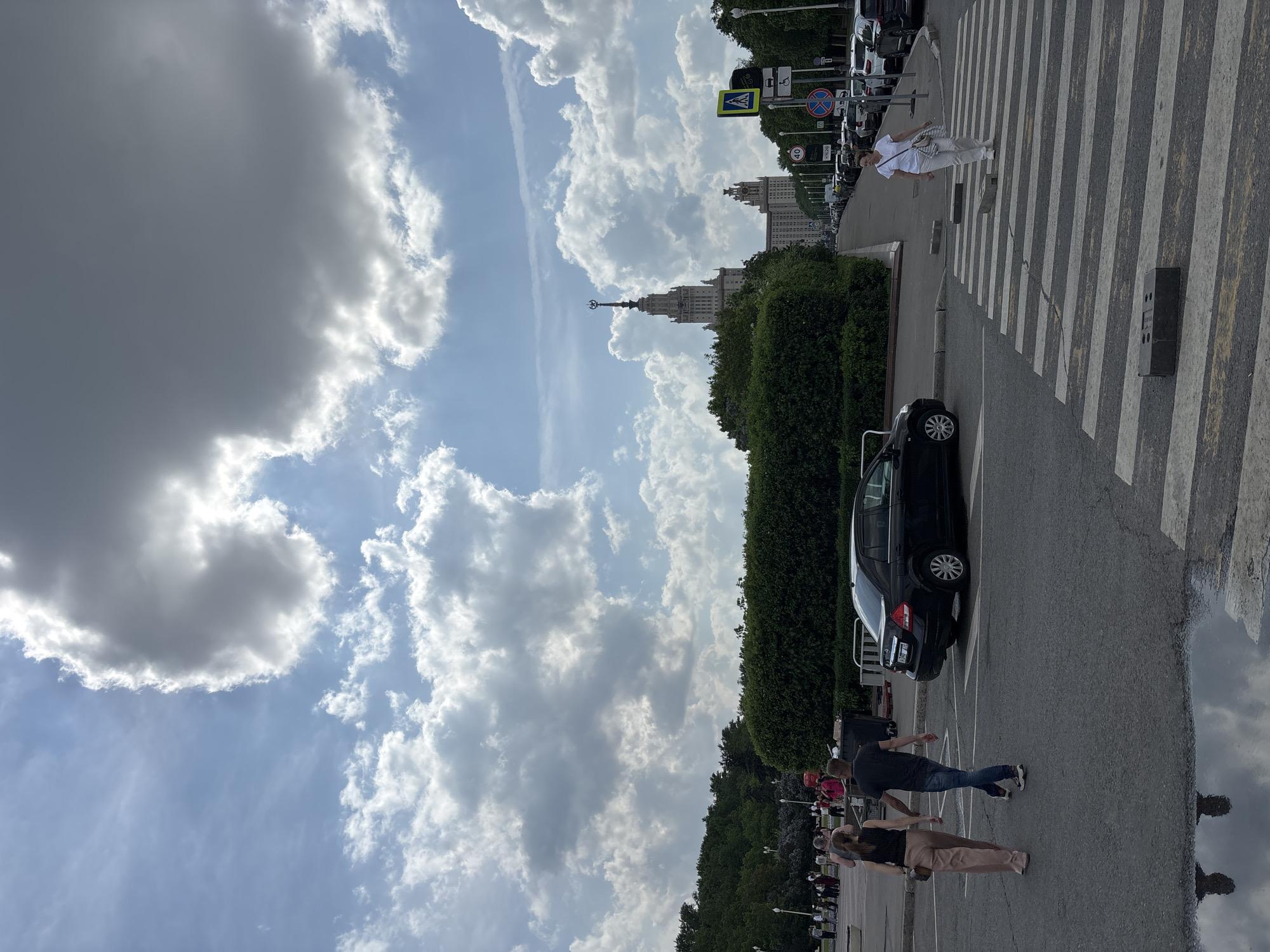 The image depicts a street scene in Moscow, Russia, likely near the Moscow State University main building, recognizable by its distinctive Stalinist architecture spire in the background. The sky is dominated by cumulus clouds. A black car is parked along the roadside. Several pedestrians are visible, some crossing the street at a crosswalk. The scene features street signs, green foliage, and a wet section of road showing a reflection.