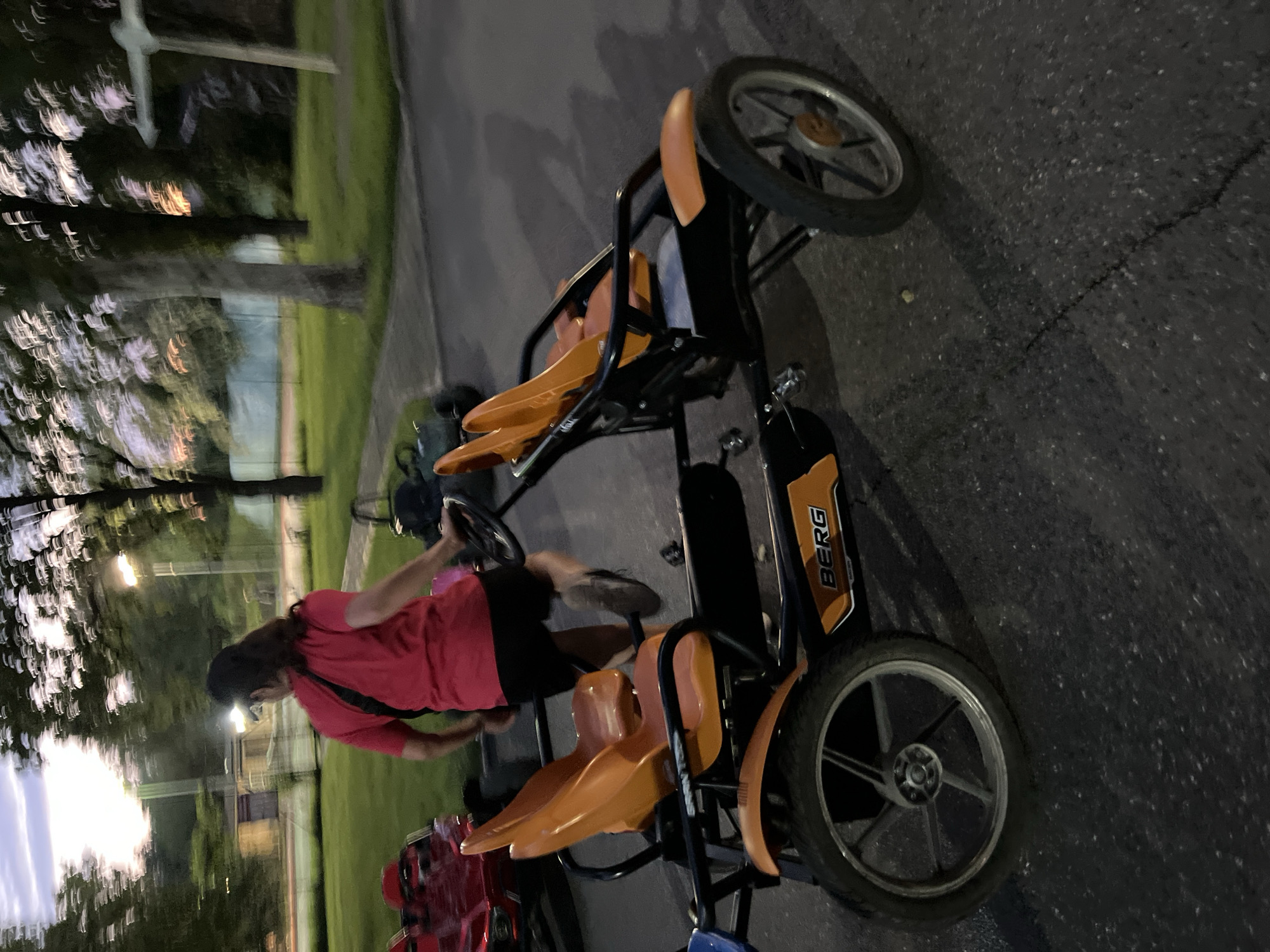 In Krasnodar, Russia, at dusk, a person wearing a red shirt and black shorts is seen from behind, standing next to a bright orange pedal go-kart. The go-kart has two seats and the word "BERG" is visible on its side. The scene appears to be an outdoor recreational area, possibly a park or playground, with trees and grassy areas in the background. The lighting suggests it is either early morning or late evening. Another red pedal go-kart is visible to the left, and a blurry green go-kart is further in the background. The ground is paved with asphalt.