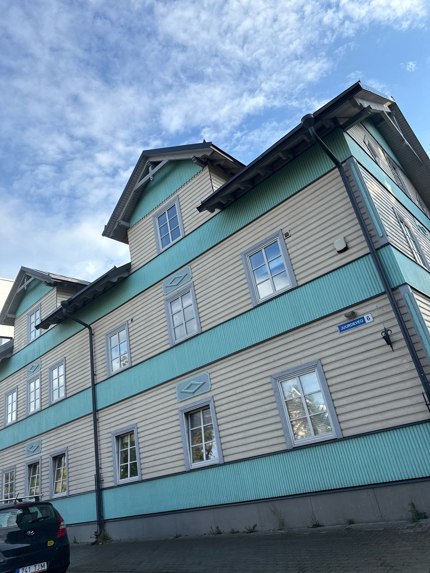 The image shows a light beige and teal multi-story building with visible wooden siding and multiple windows. The building is angled, as is the perspective of the shot, suggesting it was taken from a slightly elevated position or a tilted angle. A street sign reading "JUURDEVEÖ 6" is visible on the side of the building. A black car with a license plate is parked in the foreground. The sky is partly cloudy with a bright blue sky. The architecture suggests a European city. The building's design and the street sign are characteristic of Tallinn, Estonia.