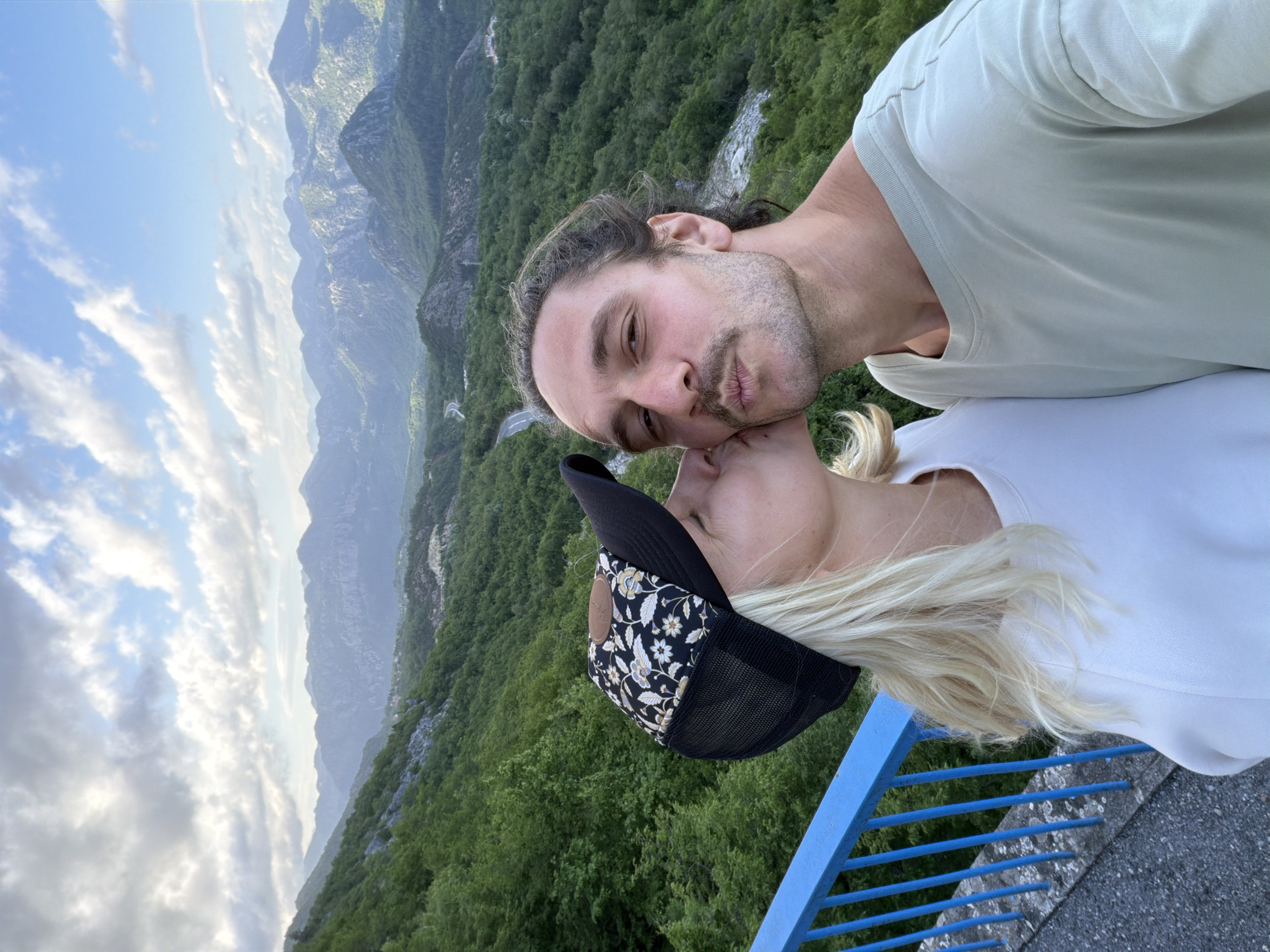 A vertically-oriented photograph captures a couple embracing closely, seemingly on an overlook in Bukovik, Montenegro. The man, with long dark hair pulled back, a mustache, and light stubble, is wearing a light green t-shirt. He has his lips pursed as if about to kiss. The woman next to him has blonde hair and wears a black baseball cap with a floral design on the front. She is wearing a white top. They are standing on what appears to be a concrete platform with a blue metal railing visible at the bottom. Behind them, a lush green mountain range extends into the distance. The sky is partially cloudy with varying shades of blue.