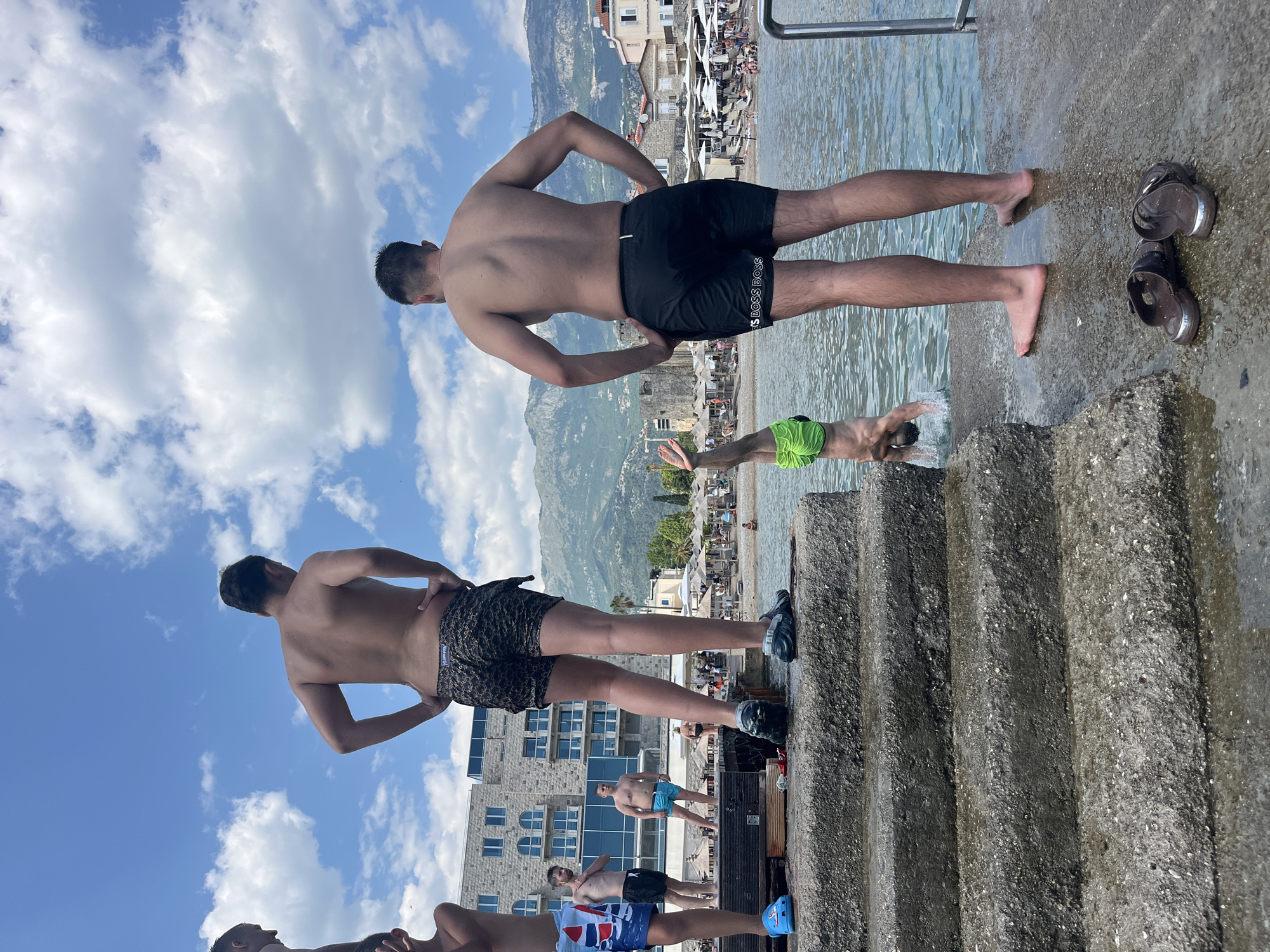 In Budva, Montenegro, on a sunny day, a group of young men are enjoying time near the Adriatic Sea. Three men are standing on concrete steps and a platform by the water, while one man is in the process of diving headfirst into the sea. The man on the far right is wearing black swim trunks with white lettering that reads "BOSS BOSS," and the man to his left is wearing patterned swim trunks. The man on the steps is wearing blue flip-flops. In the background, a beach with umbrellas and lounge chairs is visible, along with buildings and mountains. The sky is blue with scattered white clouds.