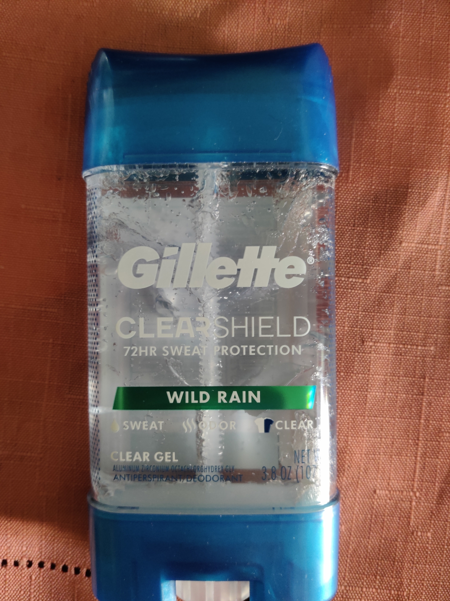 A clear antiperspirant and deodorant stick with a blue cap is positioned vertically on a rust-colored textured surface. The product is identified as "Gillette CLEAR SHIELD 72HR SWEAT PROTECTION" with a "WILD RAIN" scent. The label also indicates it is a "CLEAR GEL" and displays the net weight as "3.8 OZ (107g)". The background surface is a woven fabric with visible texture and slight shadow. There are no people or animals present in the image. The scene appears to be indoors, as suggested by the lighting and surface. No information about the specific location in Lower Paxton Township, United States of America can be derived from the image.