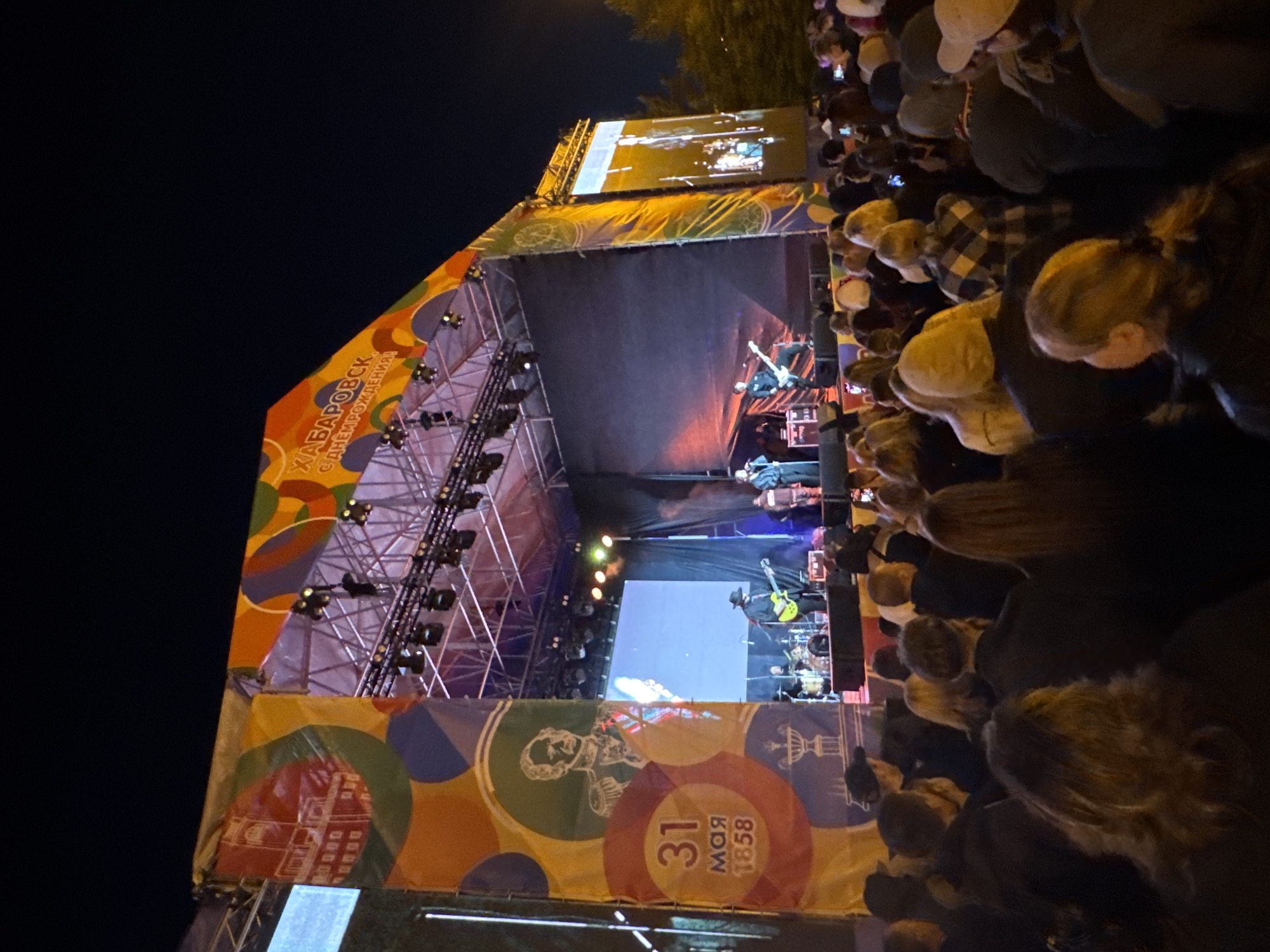At an outdoor concert in Khabarovsk, Russia, a band is performing on a large stage decorated with colorful banners. The banners feature imagery related to the city's founding, including the date May 31, 1858. The stage is equipped with lighting and a large screen above the band, displaying images. A crowd of people is gathered in front of the stage, watching the performance. The event takes place at night.