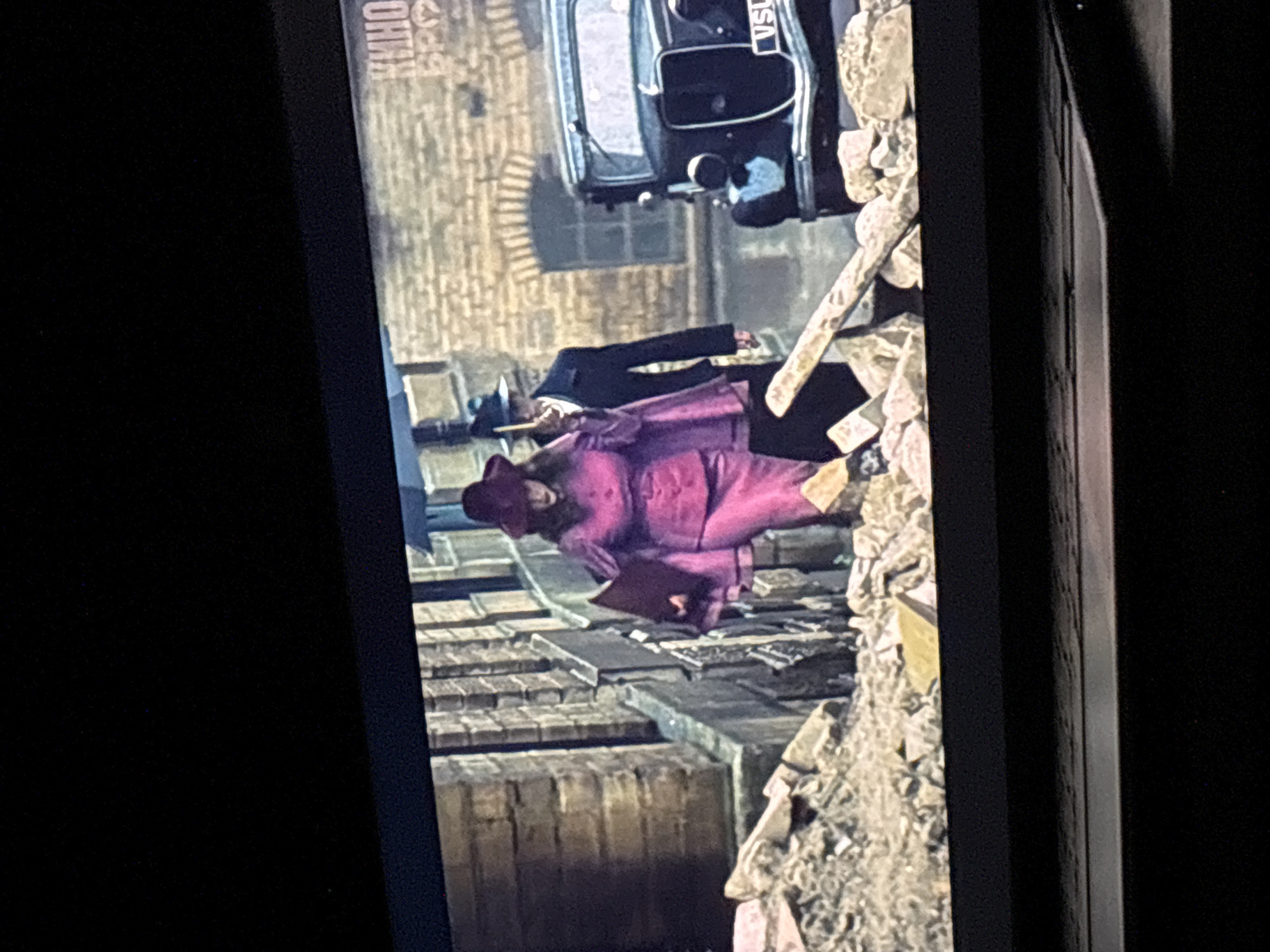 The image shows a scene from the movie "Harry Potter and the Deathly Hallows – Part 1". The scene depicts a street in what is likely London, with buildings made of brick. There is rubble on the right side of the image. A car is visible, and a woman in a pink suit is lying on the ground with another person next to her. The name "Кино Бро" is visible in the top left corner of the image. There is no evidence to suggest the location is in Budva, Montenegro.