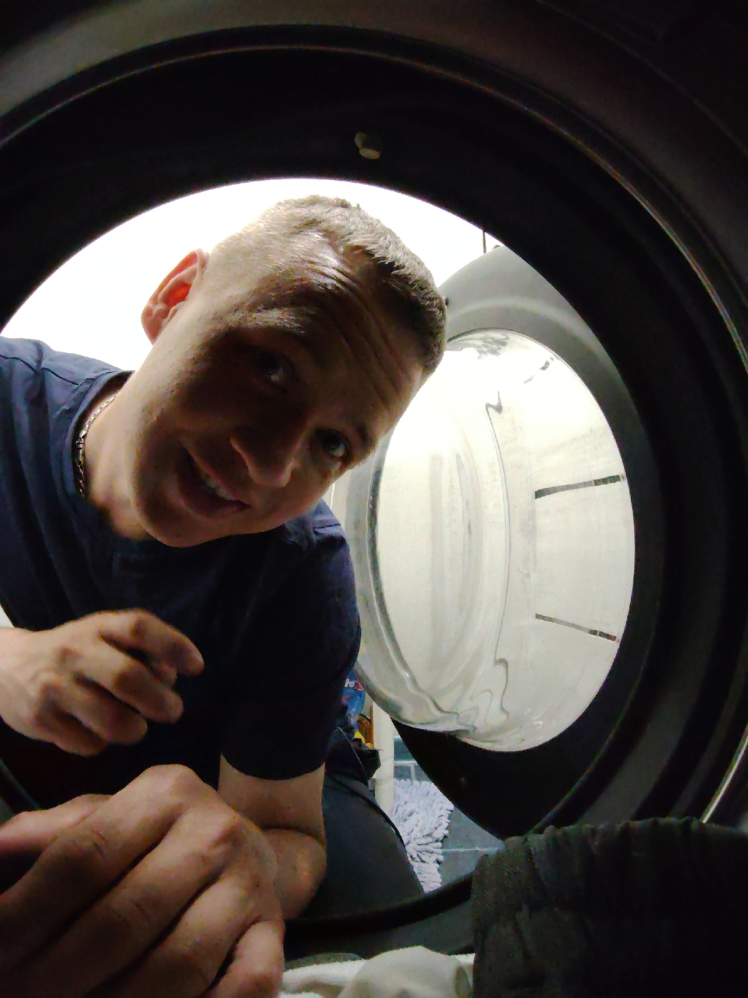 A man peers into a front-loading washing machine. The man is Caucasian, with short, light hair. He is wearing a dark t-shirt and a silver chain necklace. His face is close to the camera, and he is smiling slightly. His hand is extended towards the opening of the washing machine. Inside the washing machine, the drum is visible with some faint details. The washing machine is black and located indoors, presumably in a laundry room. In the background, a mop and some blue tiling can be seen. The location in the city of Chaikovsky, Russia is not determinable from the image.