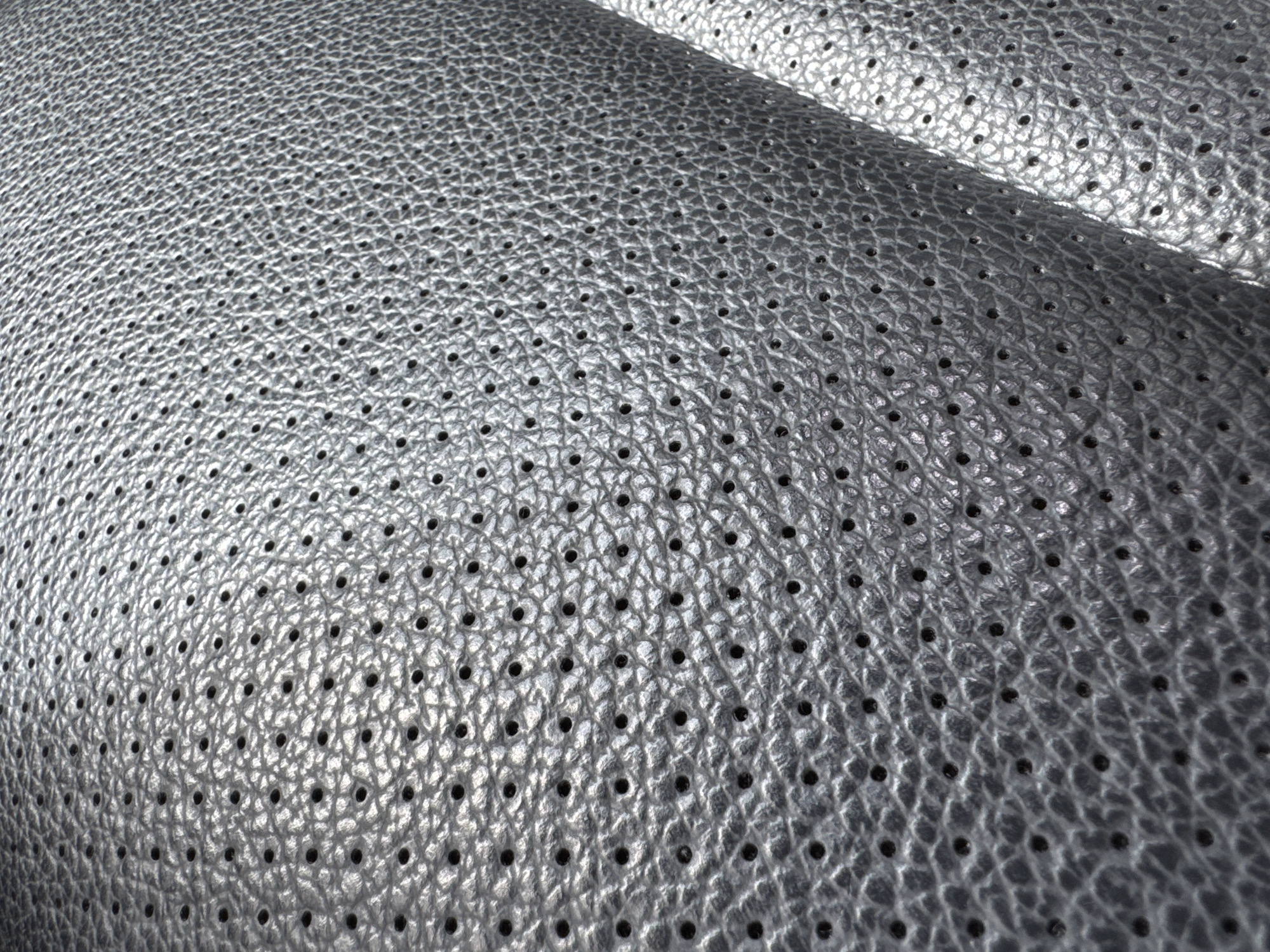 The image is a close-up, top-down view of perforated, metallic gray leather upholstery. The texture of the leather is visible, with a crinkled surface and numerous small, evenly spaced perforations. The lighting creates highlights and shadows that emphasize the three-dimensional texture of the material and the depth of the holes. The material appears to be part of a car seat or furniture. The image does not depict any elements related to the city of Folsom, United States.