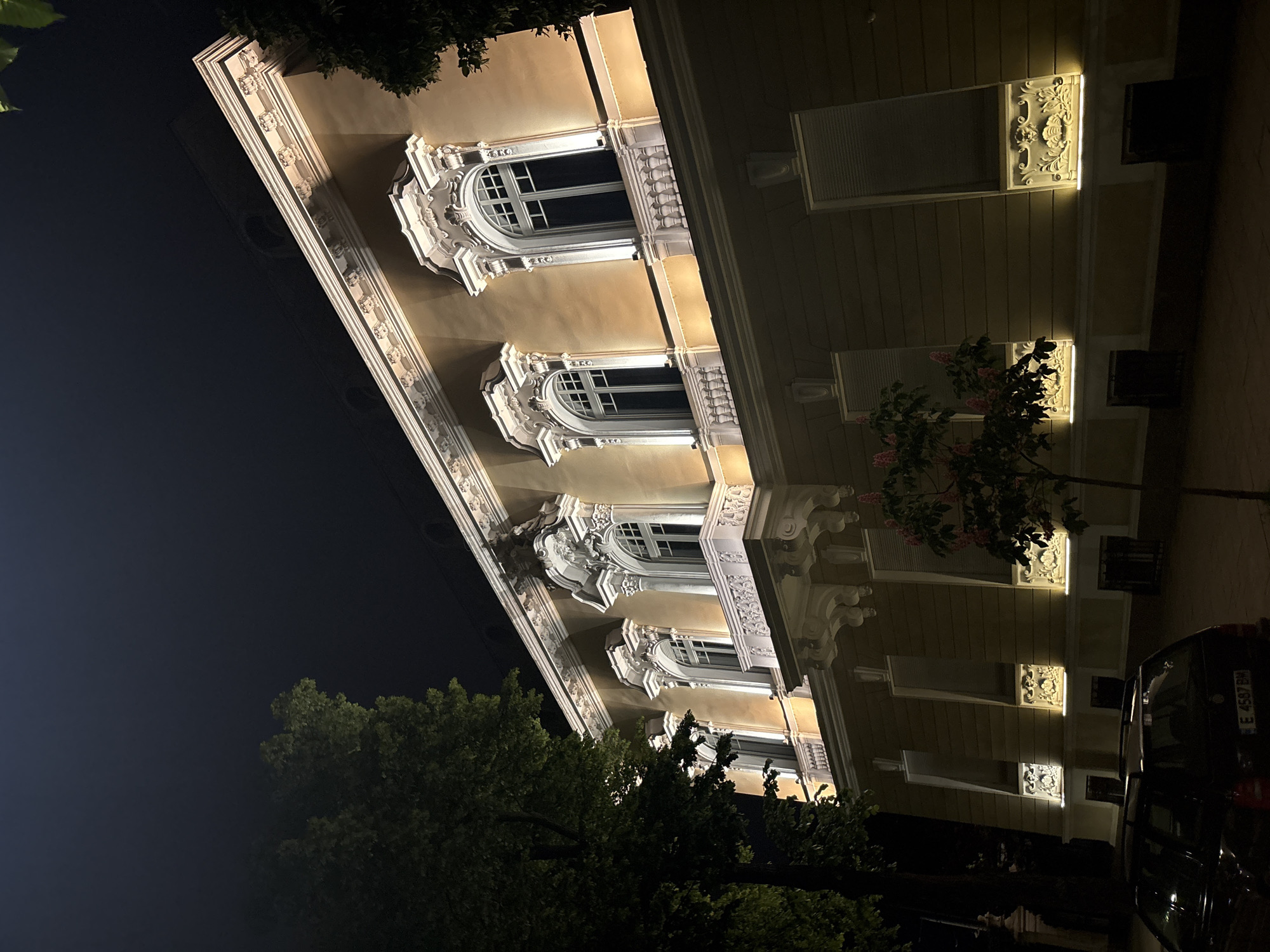 A night-time view of a building in Sofia, Bulgaria. The building is lit with yellow light and has ornate architectural details, including arched windows and decorative molding. A tree is visible in the foreground, and a dark car is parked on the street. The sky is dark.