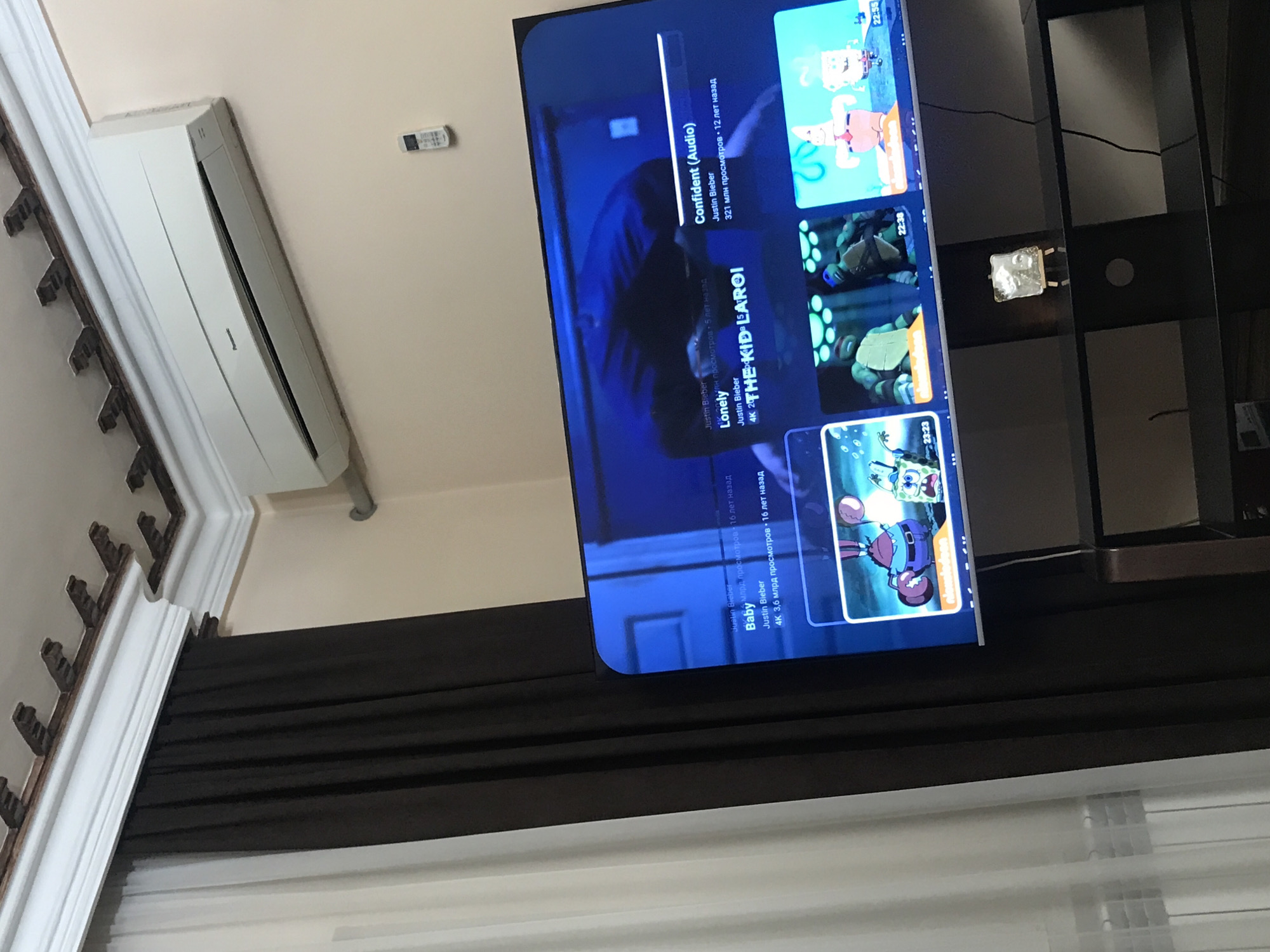 The image depicts an interior scene. A modern television screen dominates the view, displaying a YouTube interface. The displayed video suggestions include songs by Justin Bieber. A black shelving unit is visible to the right of the television. A white air conditioning unit is mounted on the wall, with a remote control situated nearby. A decorative molding runs along the top of the wall. A dark brown curtain partially obscures a window with white blinds. The image does not provide sufficient information to confirm the location as Yerevan, Armenia.