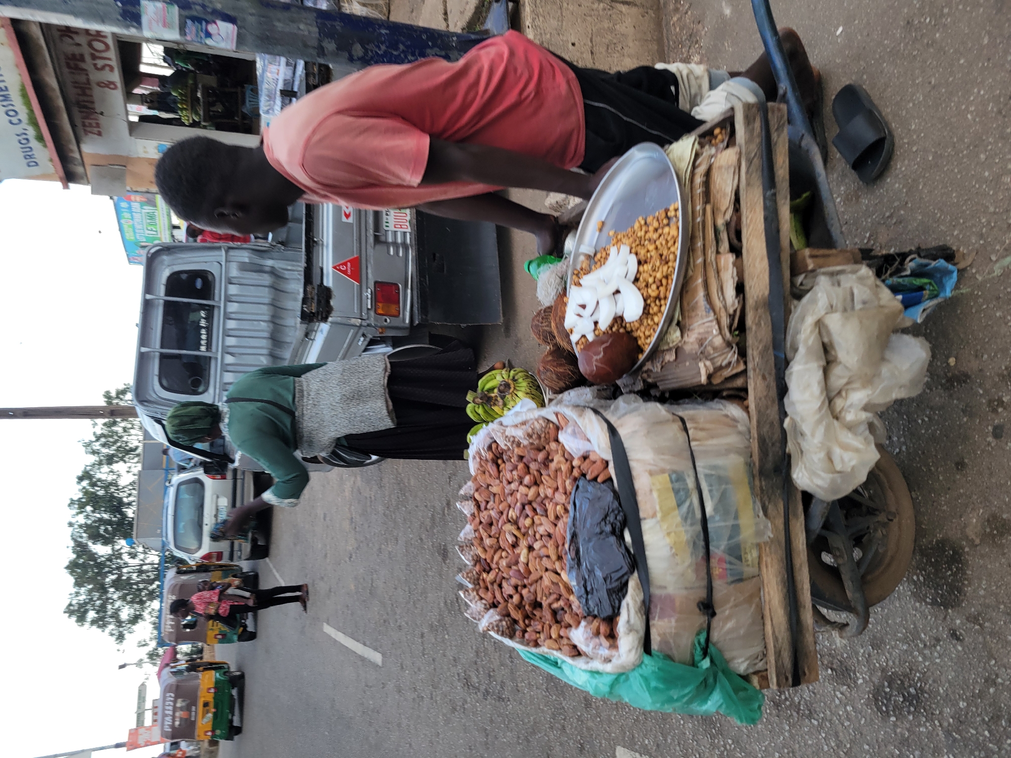 A side view of a young Black man in a pink t-shirt and black shorts is pushing a wooden cart loaded with goods. The cart contains a large pile of dried dates or similar nuts, along with some bananas and coconuts. A metal platter on the cart holds a pile of nuts, sliced white coconut pieces, and a whole coconut. The man is on a paved road in a city environment. In the background, a silver truck, several auto rickshaws, and a building with signage that reads "DRUGS, COSMETICS ZENITHLIFE PH & STORES" are visible. The scene appears to be a street market or roadside vendor location in Jos, Nigeria. The overall atmosphere suggests a busy, commercial street.